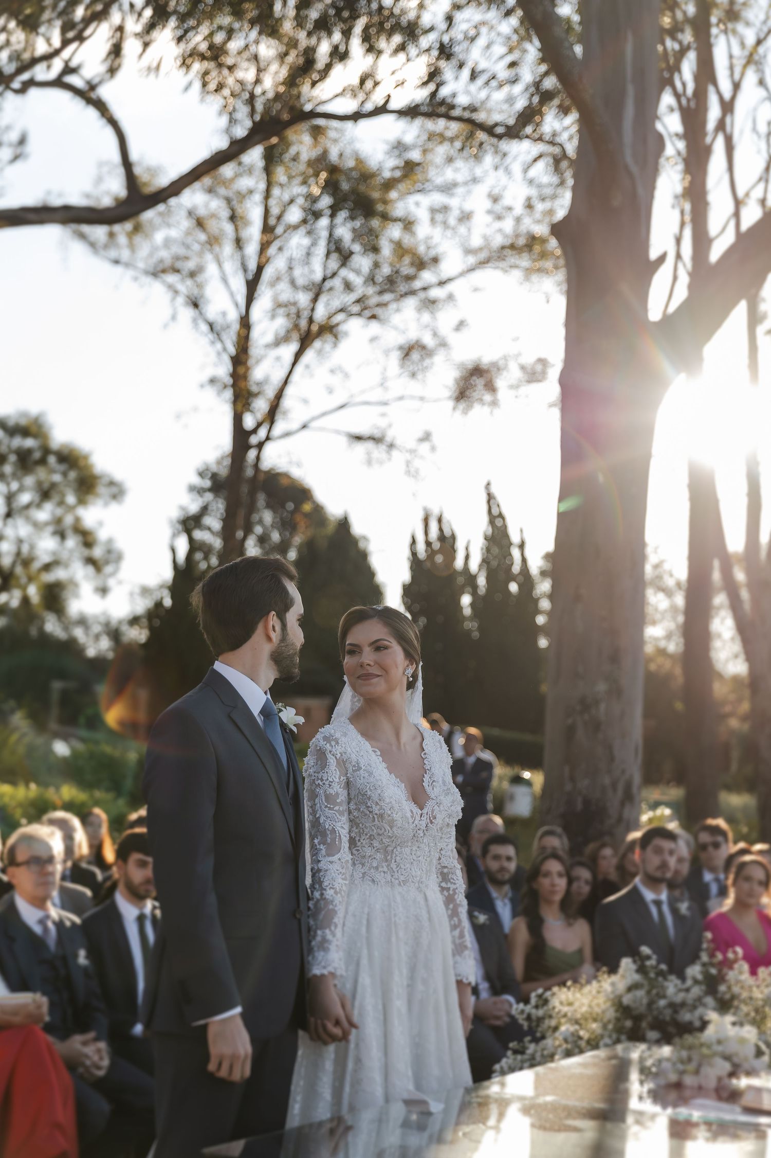 Wedding ceremony taking place outdoors at sunset with guests seated under tall trees.