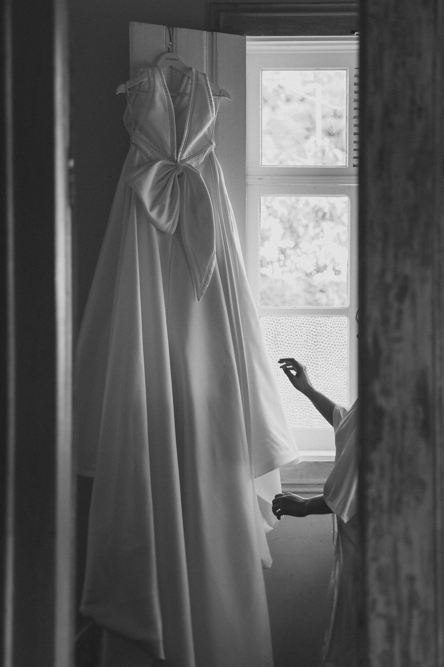 A wedding dress hanging by a window in black and white, with an arm reaching out from behind a curtain.