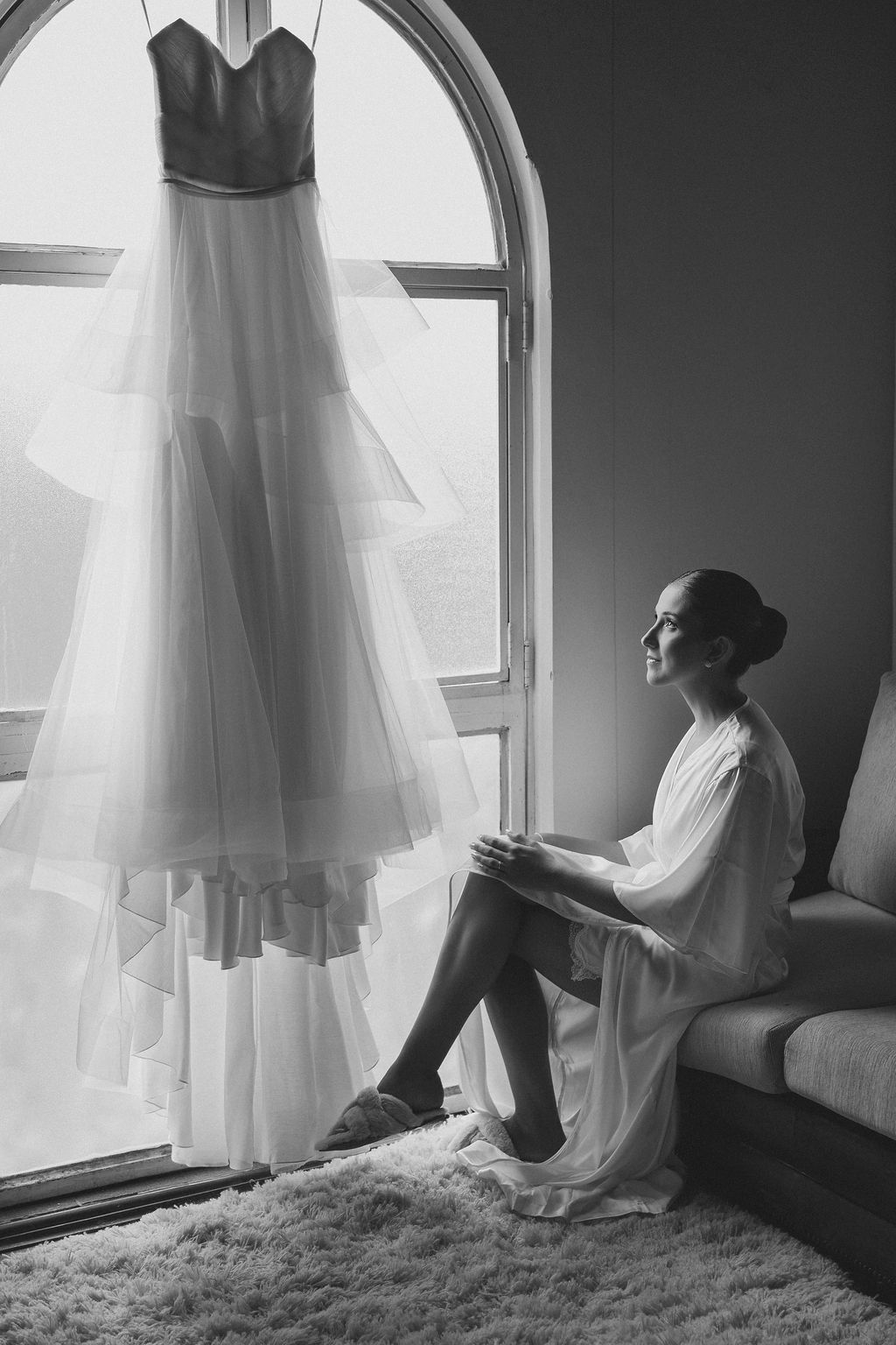 Cerimônia de casamentoA black and white photo showing an elegant wedding dress hanging by an arched window as someone sits nearby in a robe.