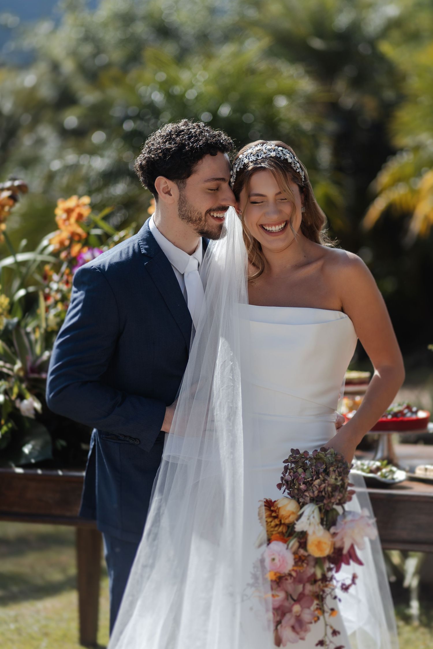 Newlywed couple shares joyful moments in sunlit garden setting with bride carrying colorful autumn bouquet.