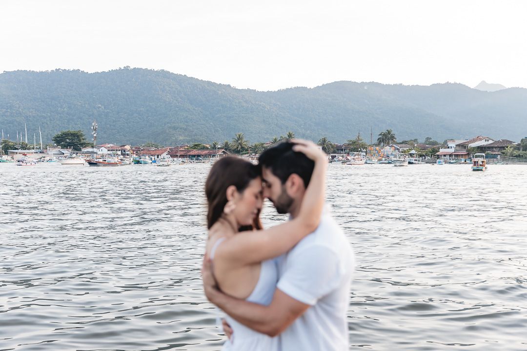 A romantic couple embraces by the waterfront with mountains in the background during sunset.