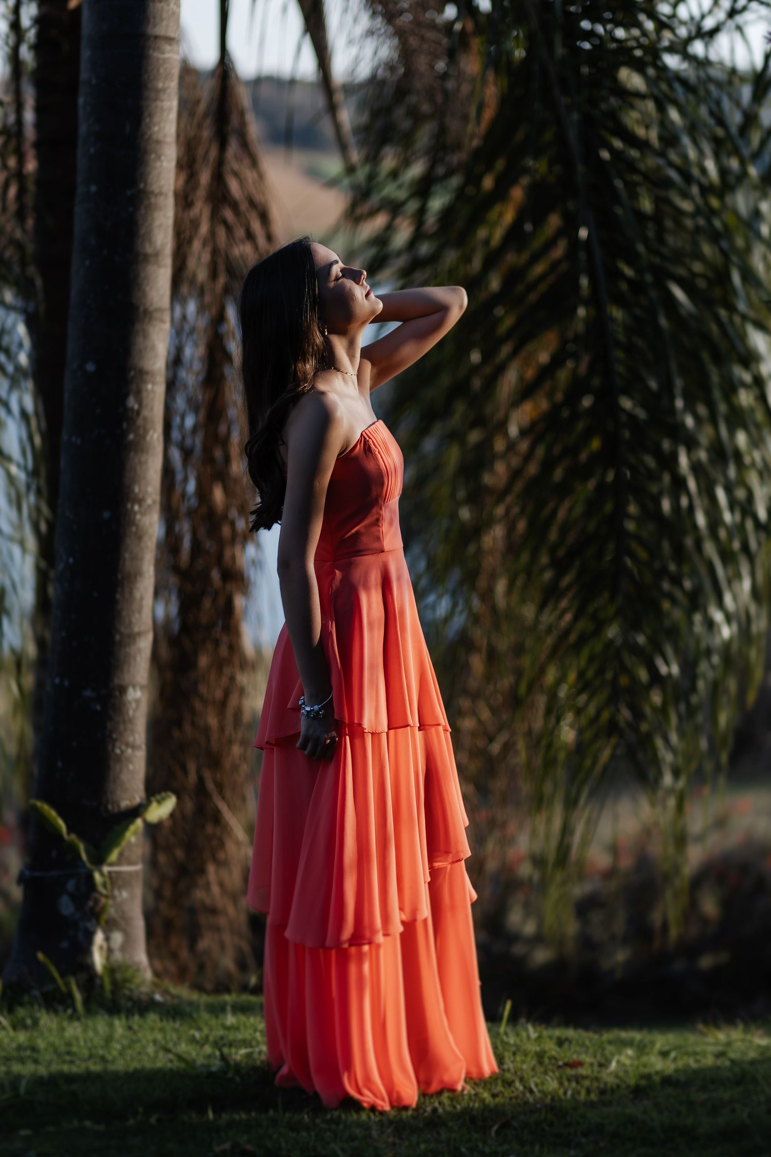 A person in a flowing coral tiered maxi dress poses gracefully among palm trees in warm sunset lighting.
