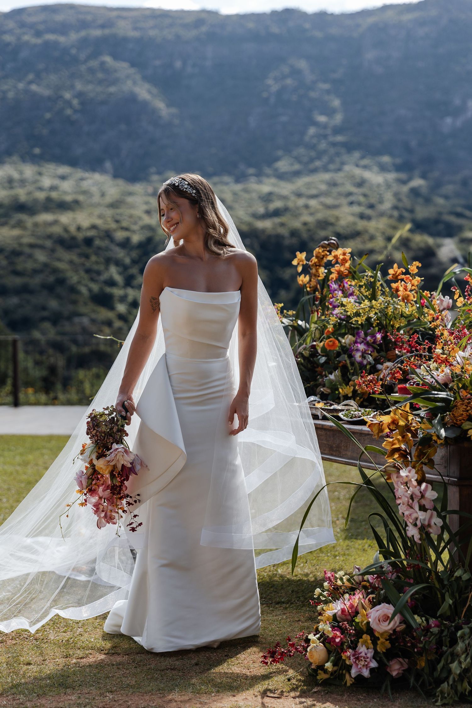 A bride in an elegant white strapless gown and flowing veil stands among colorful floral arrangements on a mountainside.
