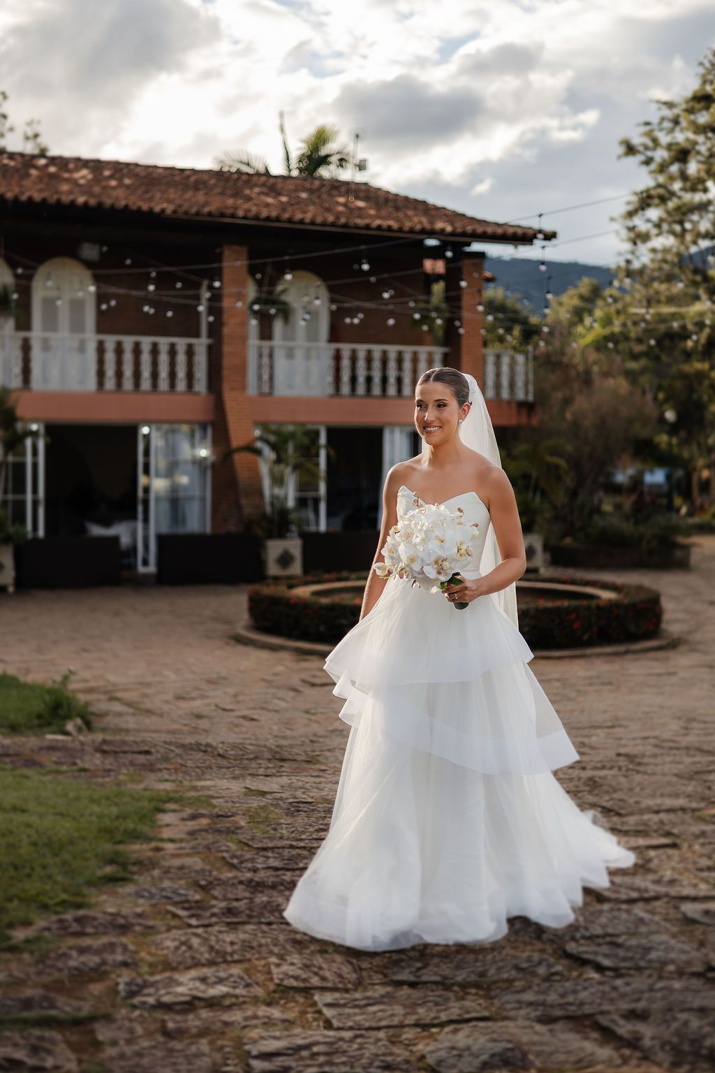 Bride in layered white wedding gown stands on stone patio with Spanish colonial style building in background.