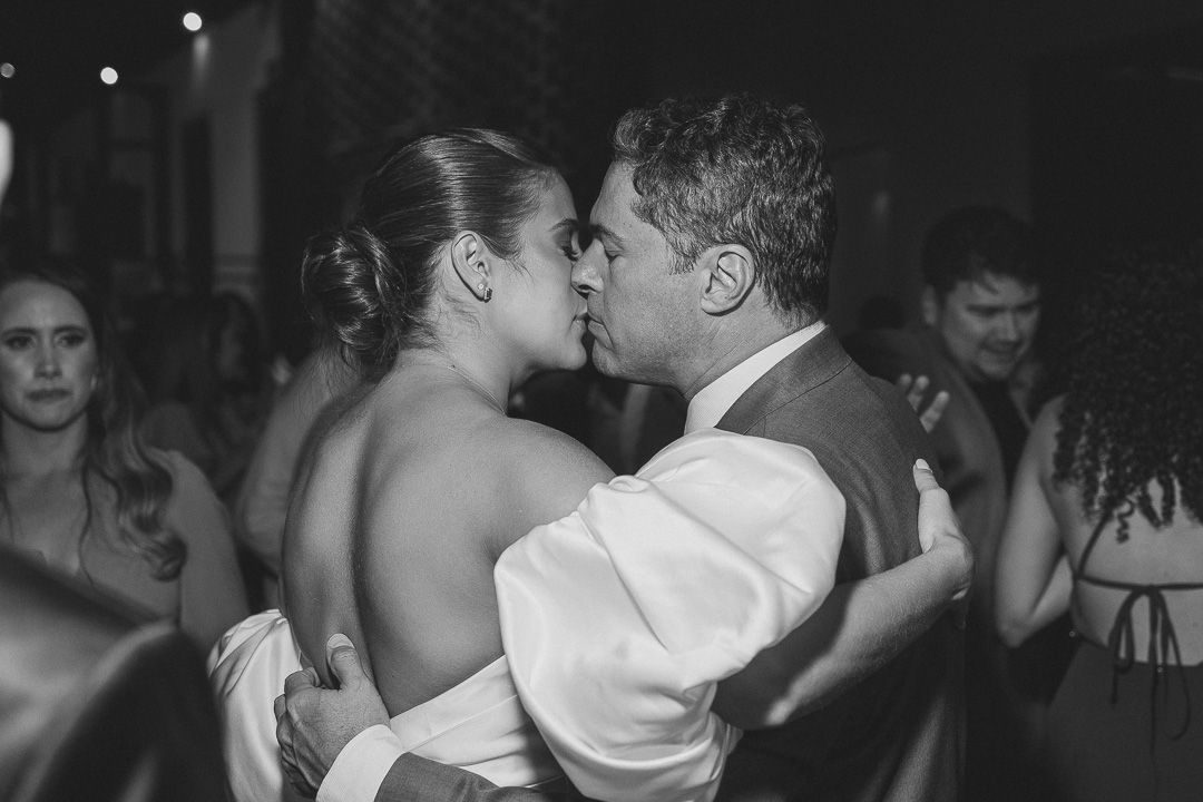 A couple shares an intimate kiss on the dance floor at a reception in a black and white candid photograph.