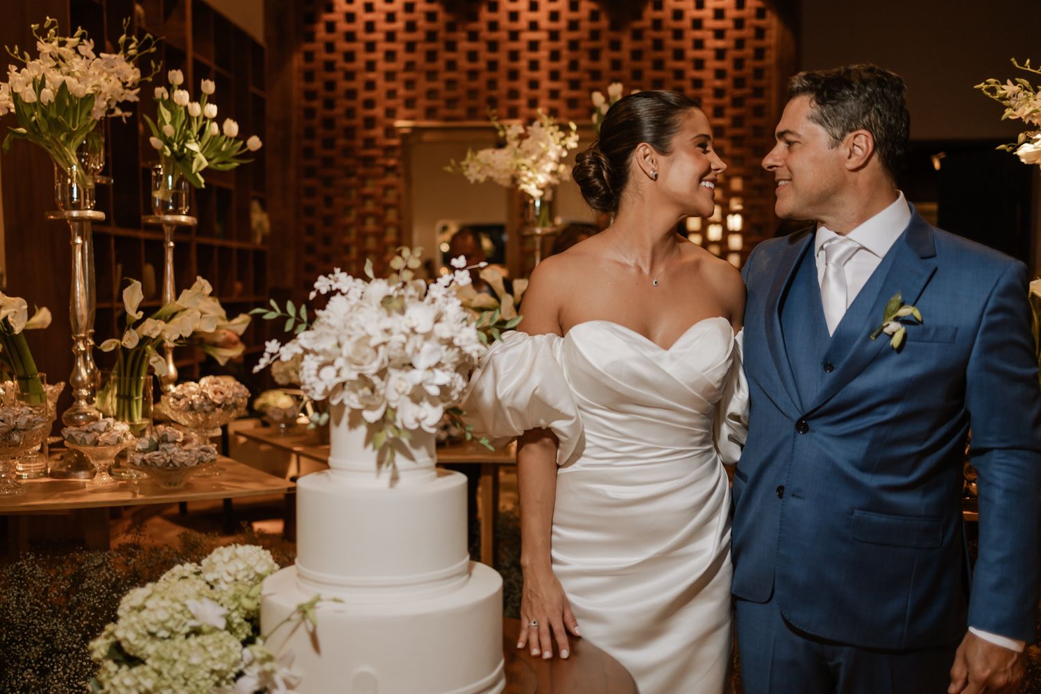A couple gazes at each other beside their elegant white wedding cake in a warmly lit reception venue.