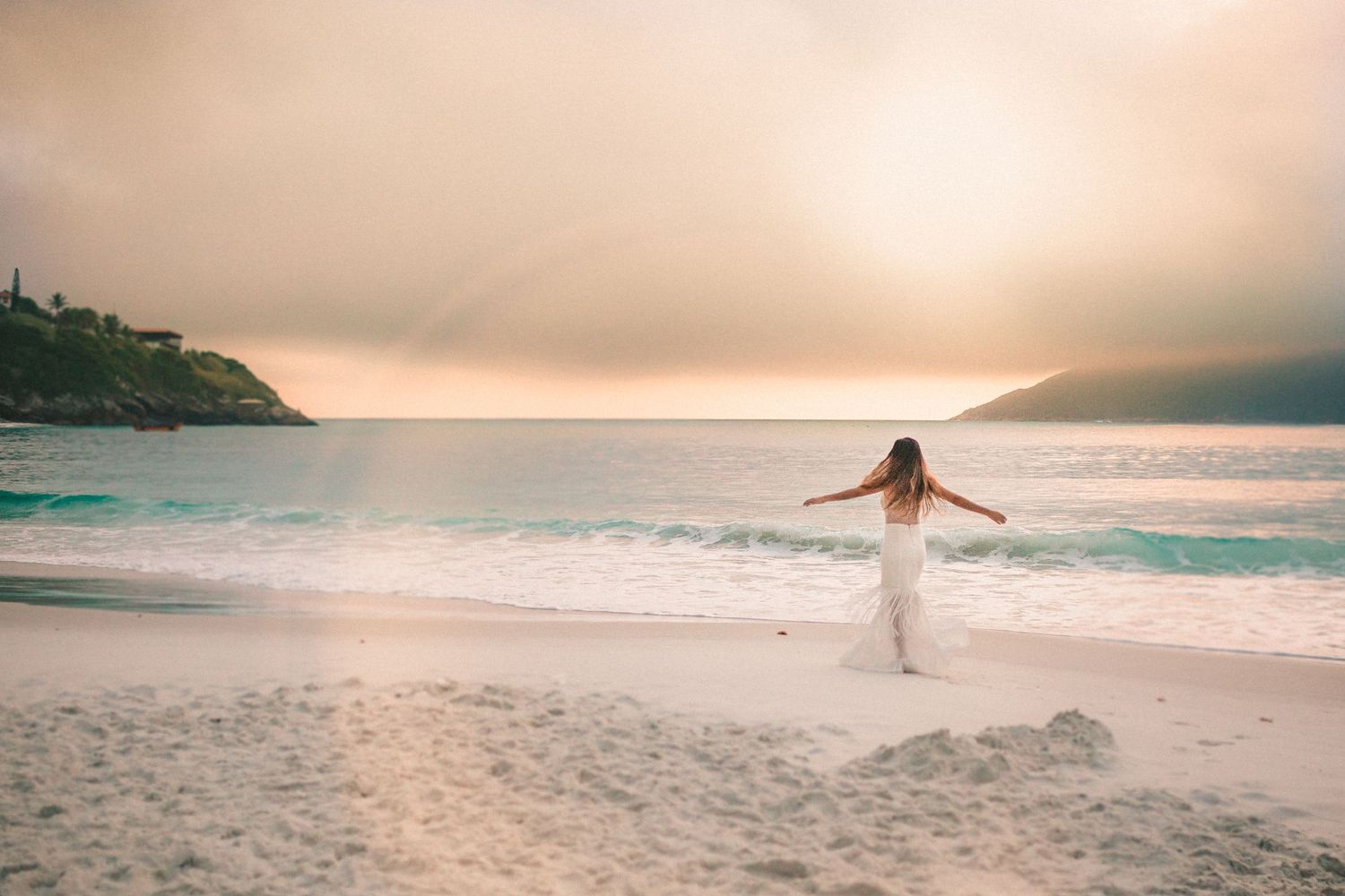 A silhouette on a sandy beach spreads their arms wide against a misty sunset over turquoise ocean waves.