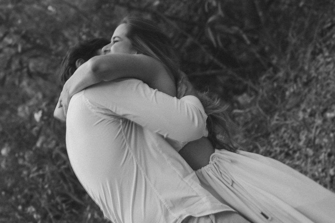 A couple in white clothing embraces while lying on grass in this tender black and white photograph.