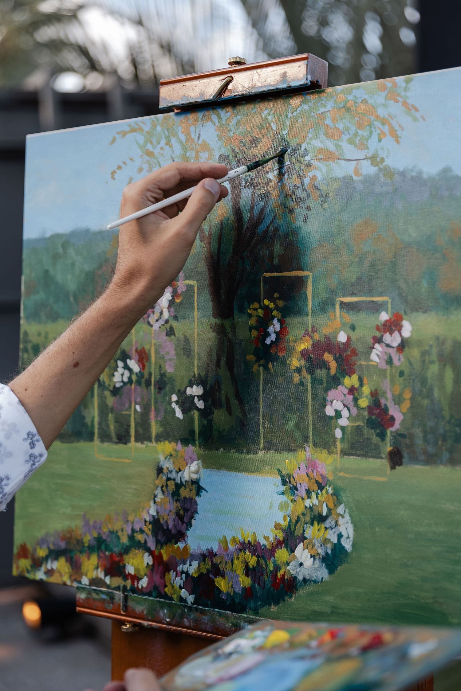 An artist's hand paints colorful flowers on a canvas depicting a garden landscape with vibrant floral arches.