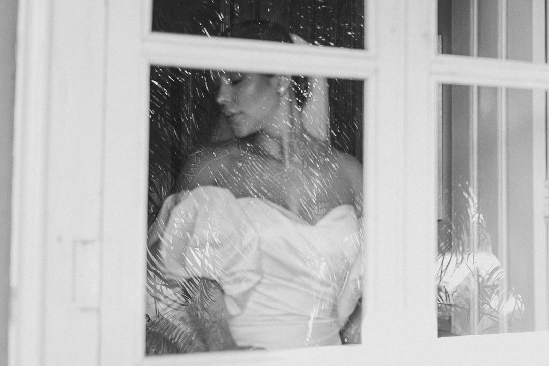 Black and white artistic photograph showing a person in a white strapless gown through a window pane.