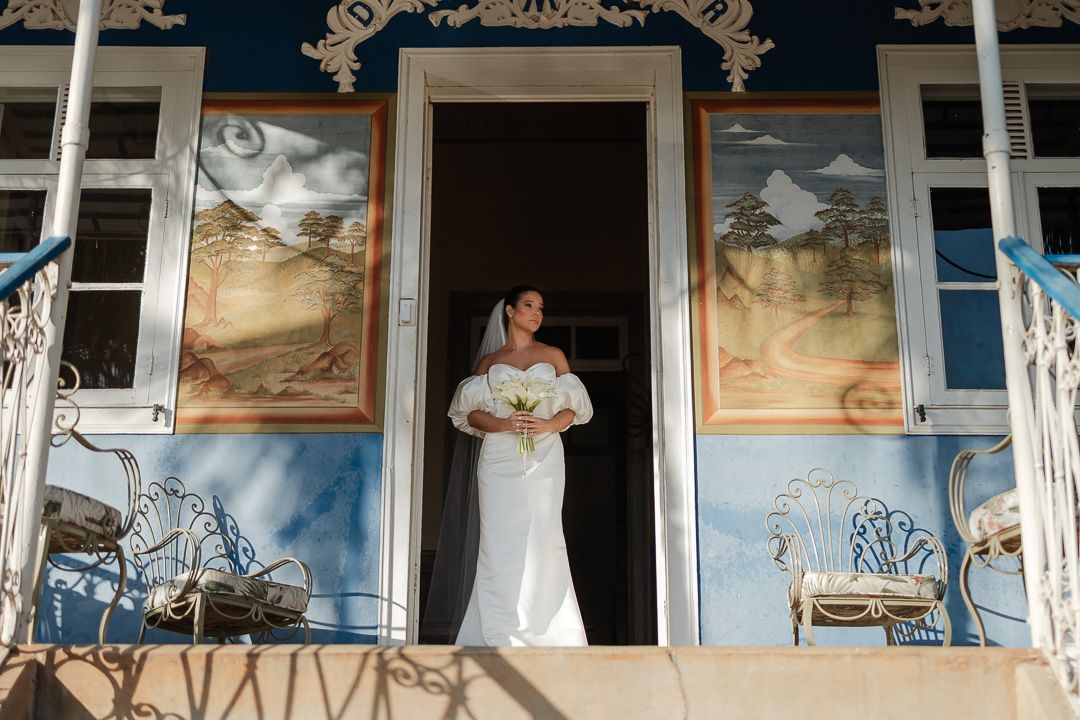 A bride in a white dress stands in a doorway flanked by ornate blue walls with decorative paintings and vintage furniture.