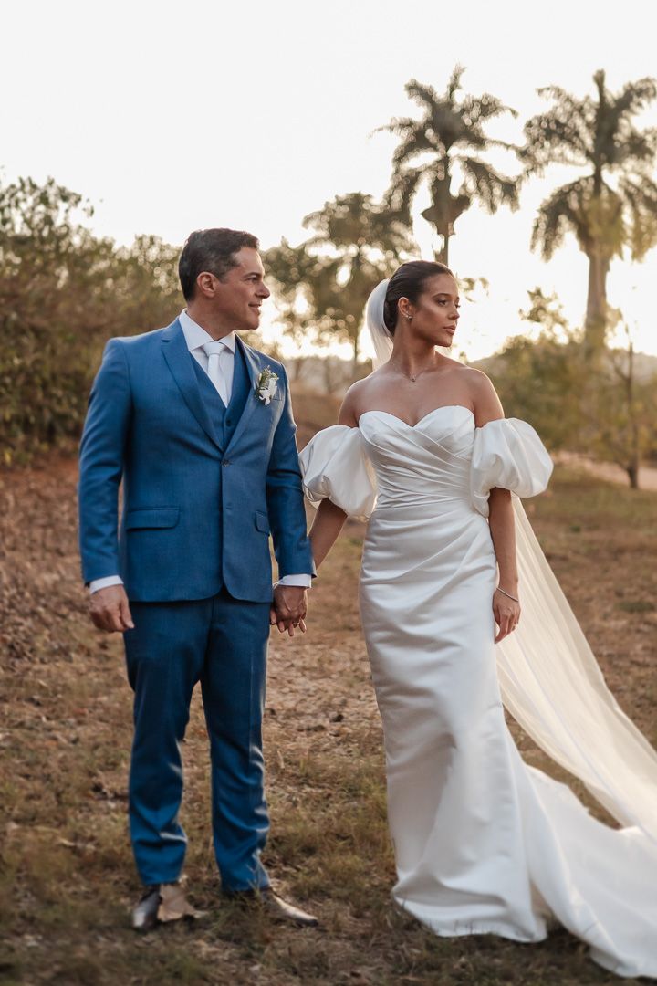 A bride in a white gown with puff sleeves and groom in blue suit pose at sunset with palm trees in the background.