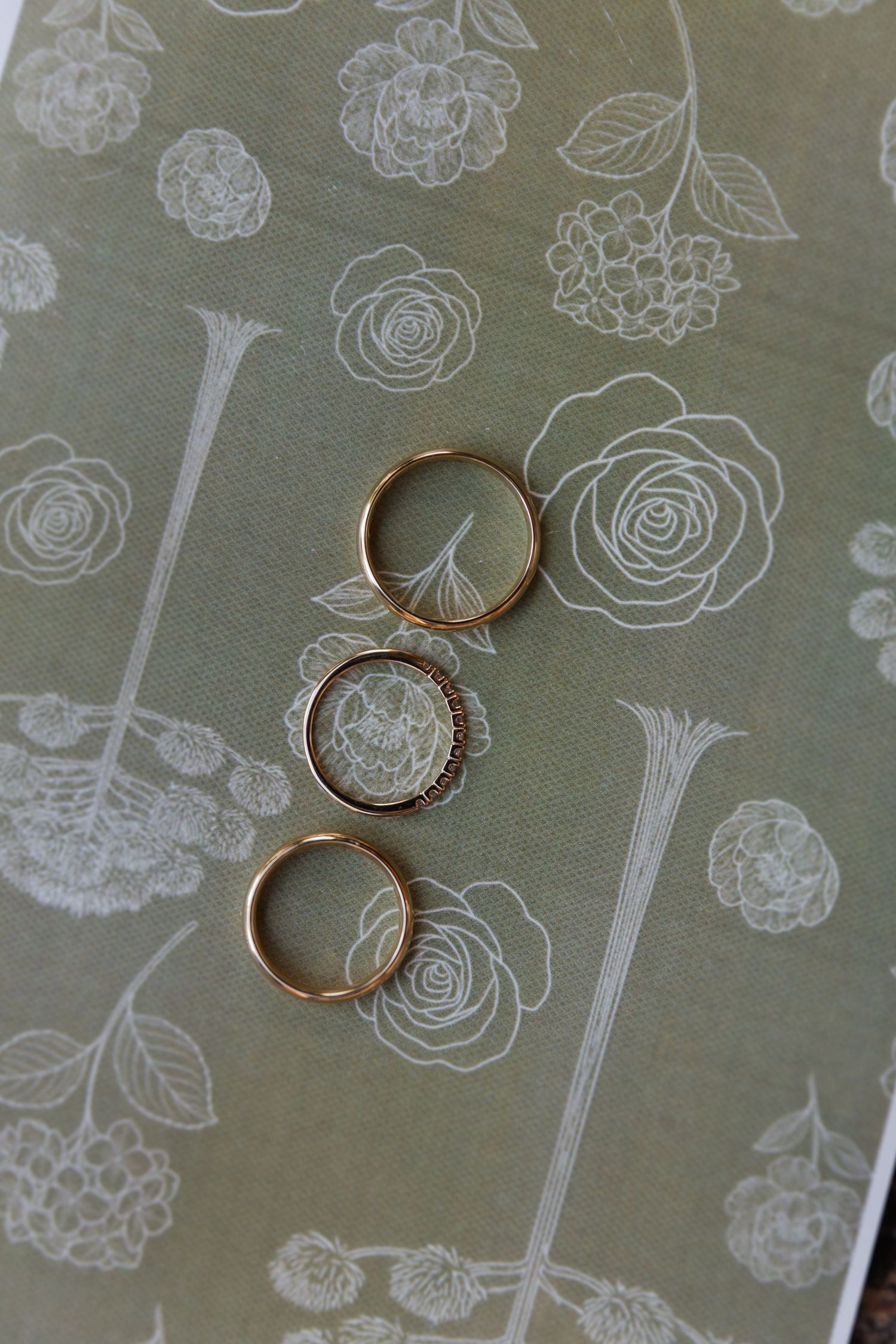 Gold wedding rings rest on a sage green fabric with delicate white floral pattern details.