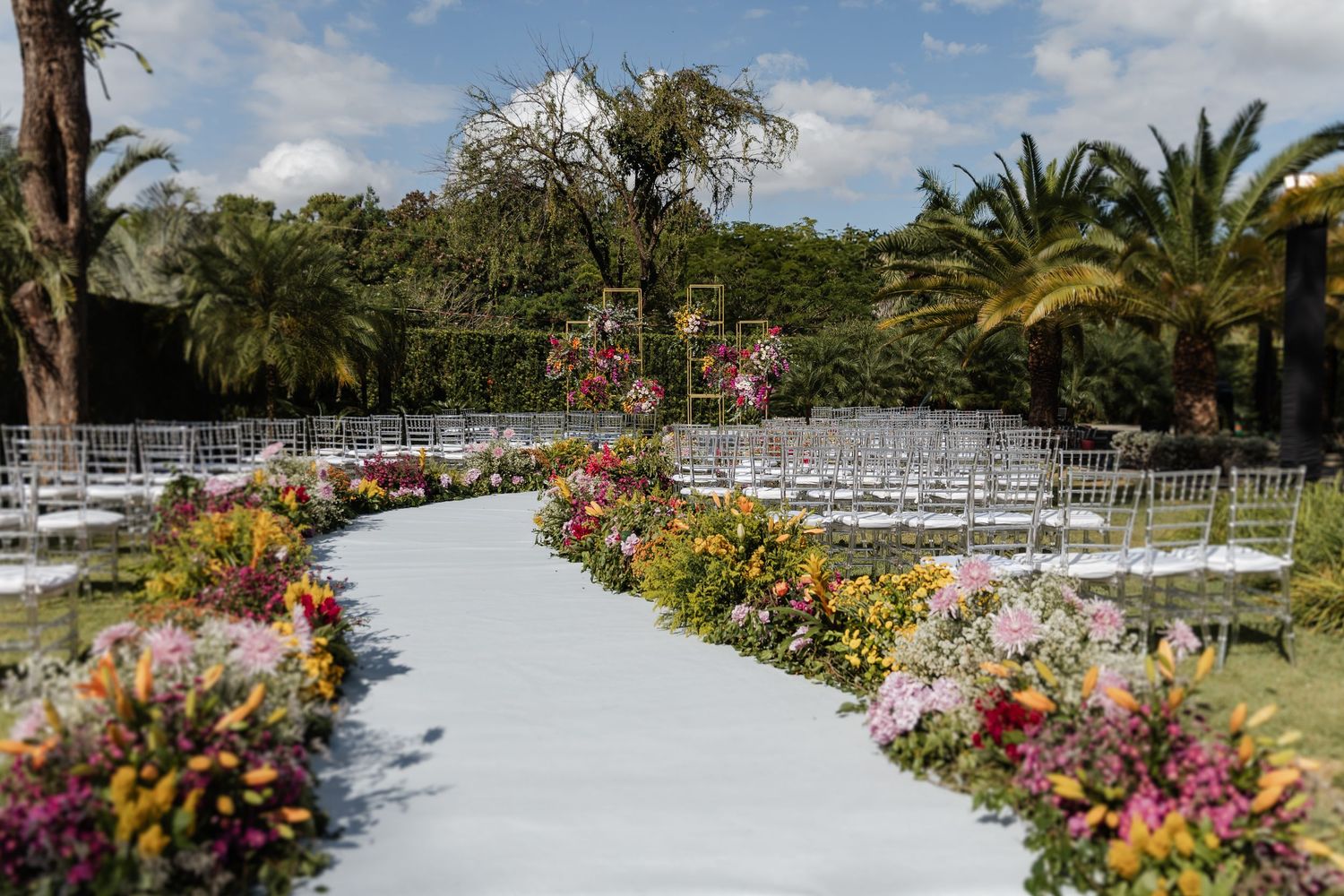 casamento em Belo Horizonte, A white curved aisle lined with colorful flowers leads through palm trees to an outdoor wedding ceremony setup.