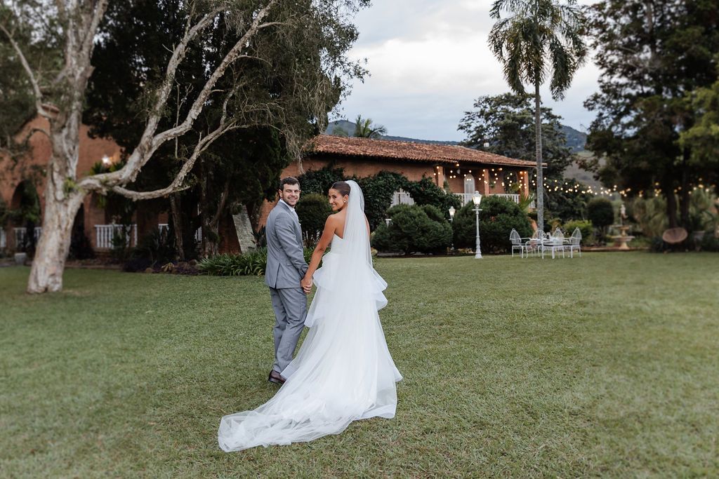 A couple in wedding attire stands on a lush green lawn in front of a Spanish-style building with terra cotta roof tiles.