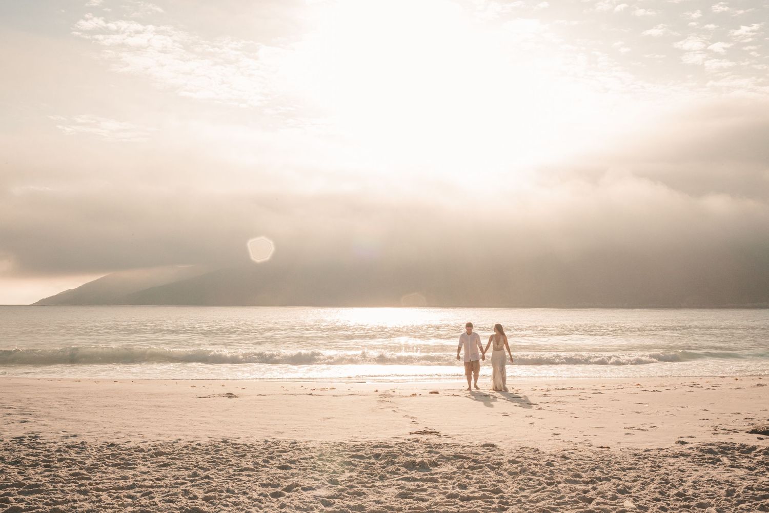 Two silhouettes walking hand in hand on a misty beach at sunset with mountains in the distance and sun glowing through clouds.