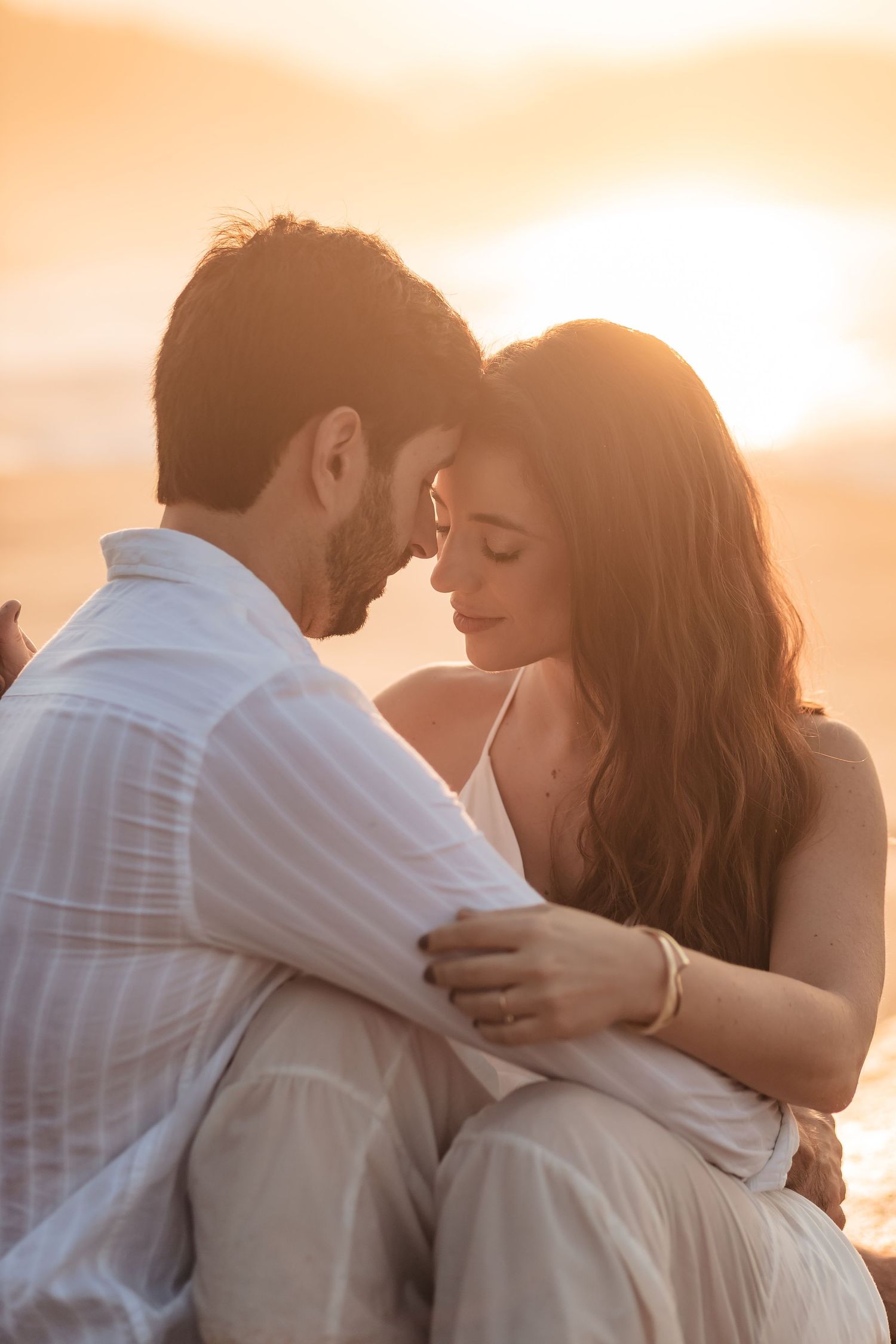 Romantic couple in white outfits embrace during golden hour sunset creating an intimate silhouette against warm sky.