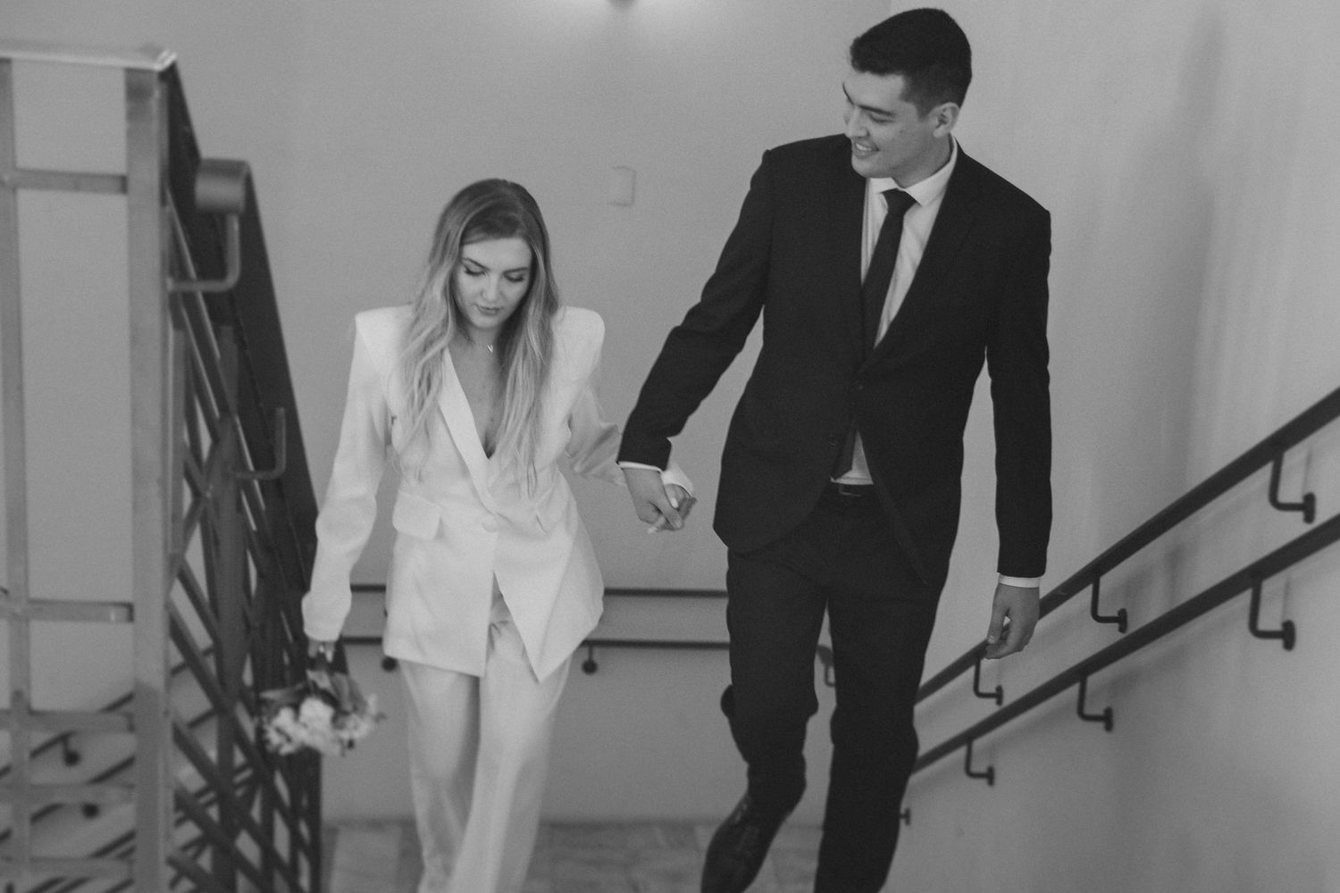 A couple in formal attire descends a staircase while holding hands in a black and white photo.