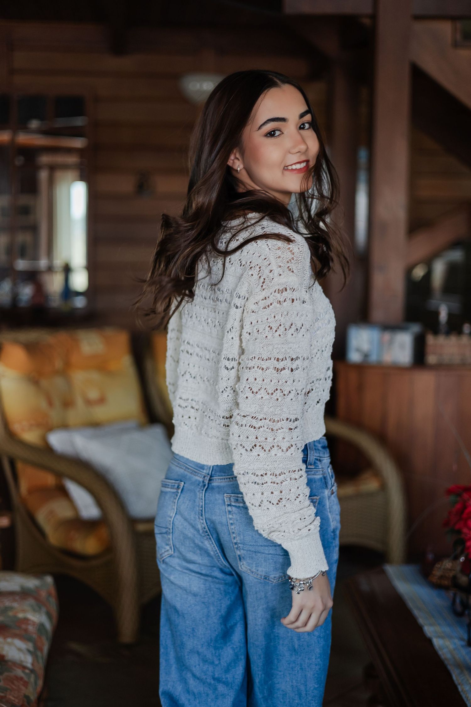 Woman wearing a white crochet sweater and high-waisted jeans poses in a rustic interior setting.
