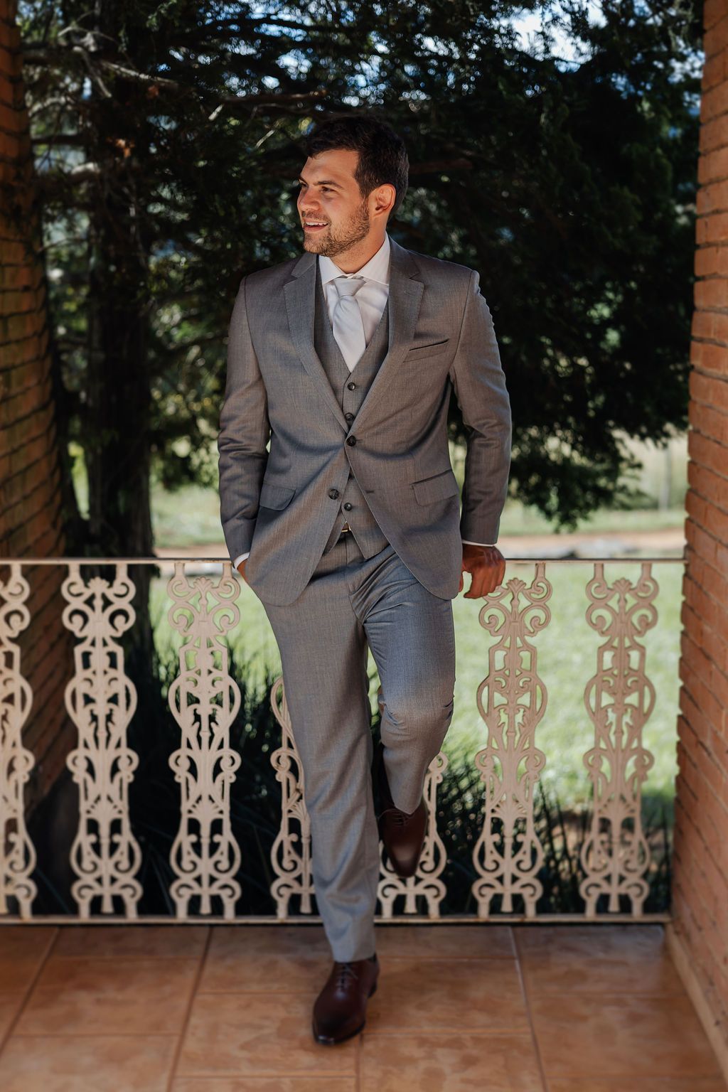 Well-dressed person in a gray suit and tie leans against ornate railing on a covered porch with trees in background.
