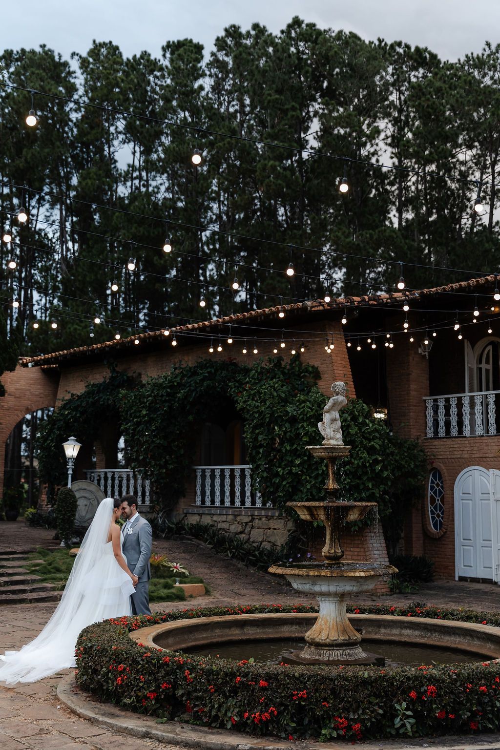 A wedding couple stands near an elegant fountain at a Spanish-style venue with string lights and tall pine trees at dusk.