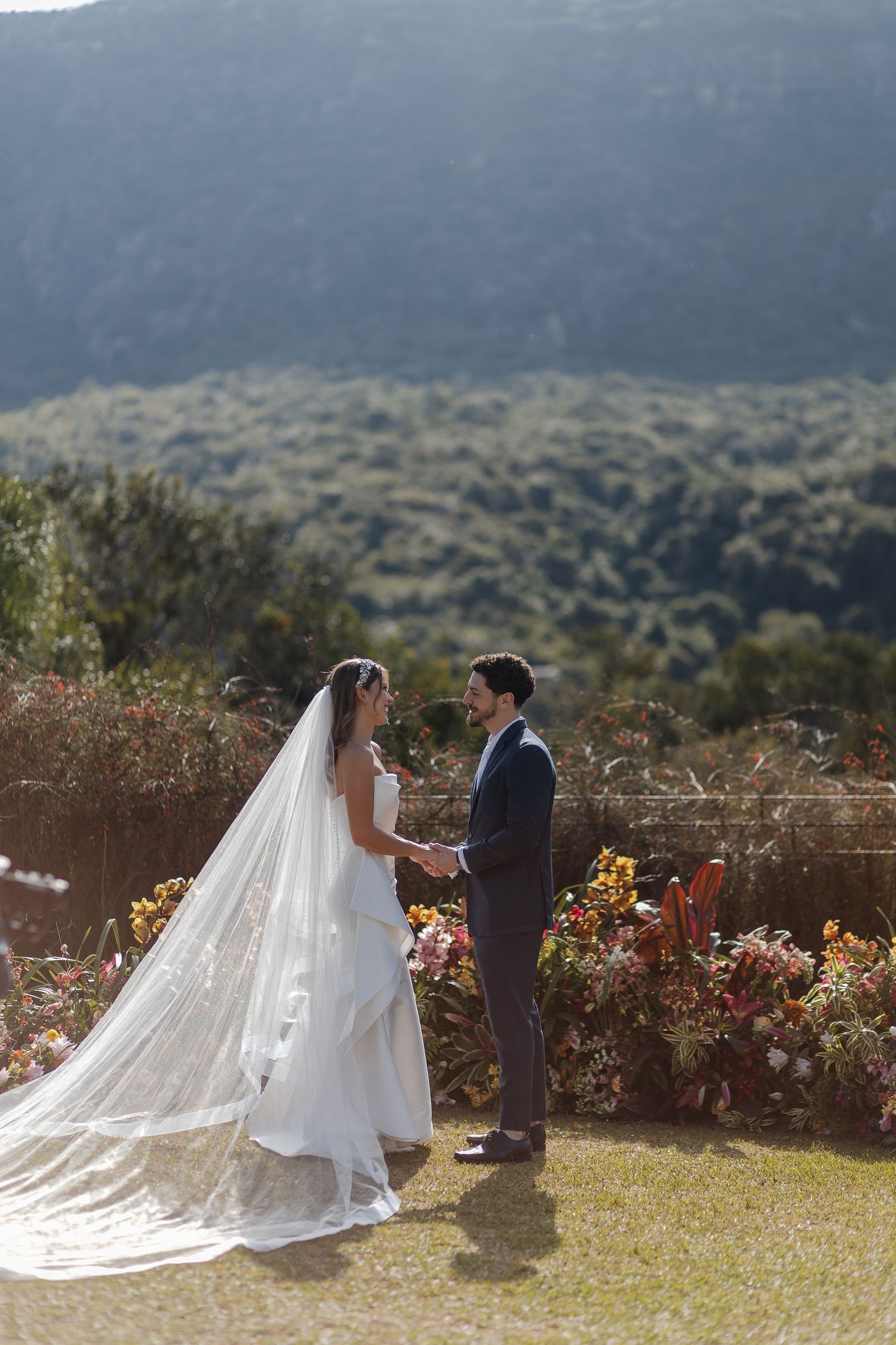A couple shares a romantic moment during their outdoor wedding ceremony against a mountain backdrop with flowing veil and flowers.