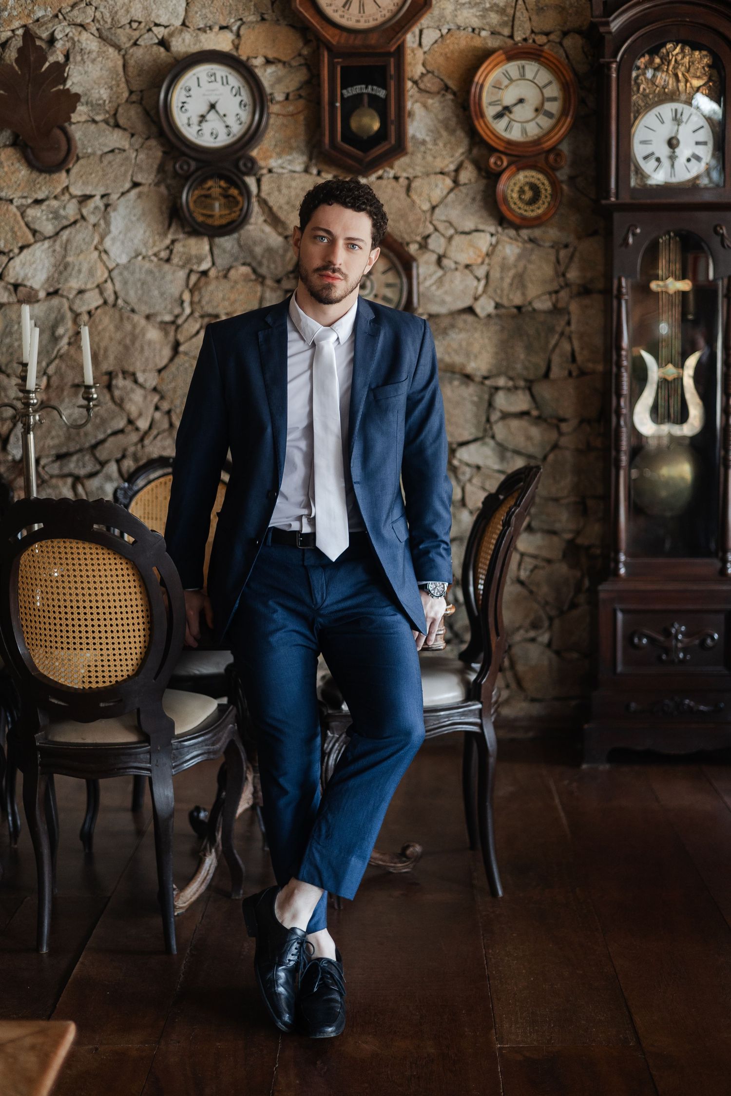 A groom in a navy suit sits in a dimly lit room with vintage clocks on stone walls creating an antique atmosphere.