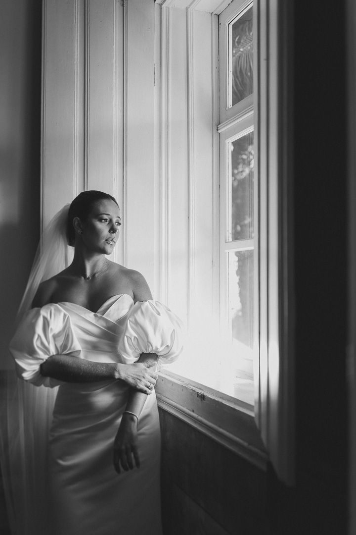 A bride in a white strapless gown with puff sleeves gazes thoughtfully out a window in black and white.