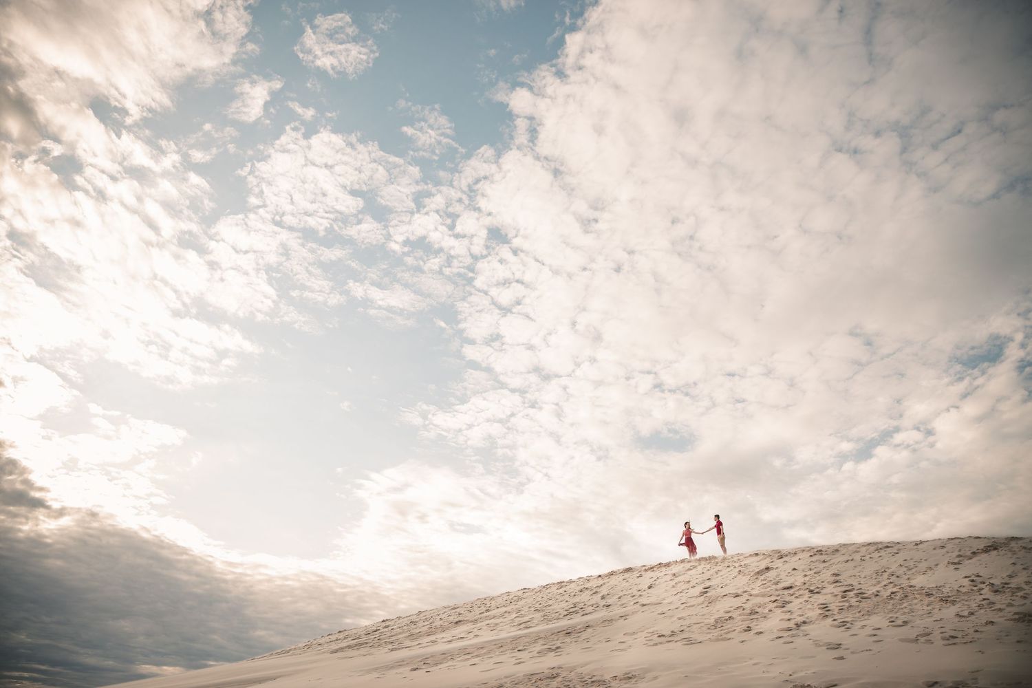 Two silhouetted figures stand atop a sandy dune against a dramatic cloudy sky at sunset.