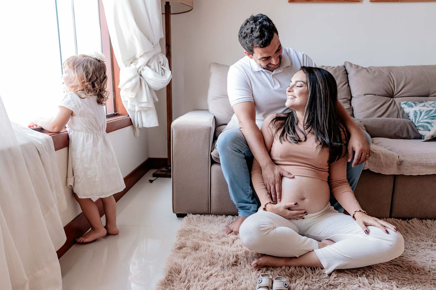 Expectant parents share an intimate moment in their living room while a young child plays by the window.