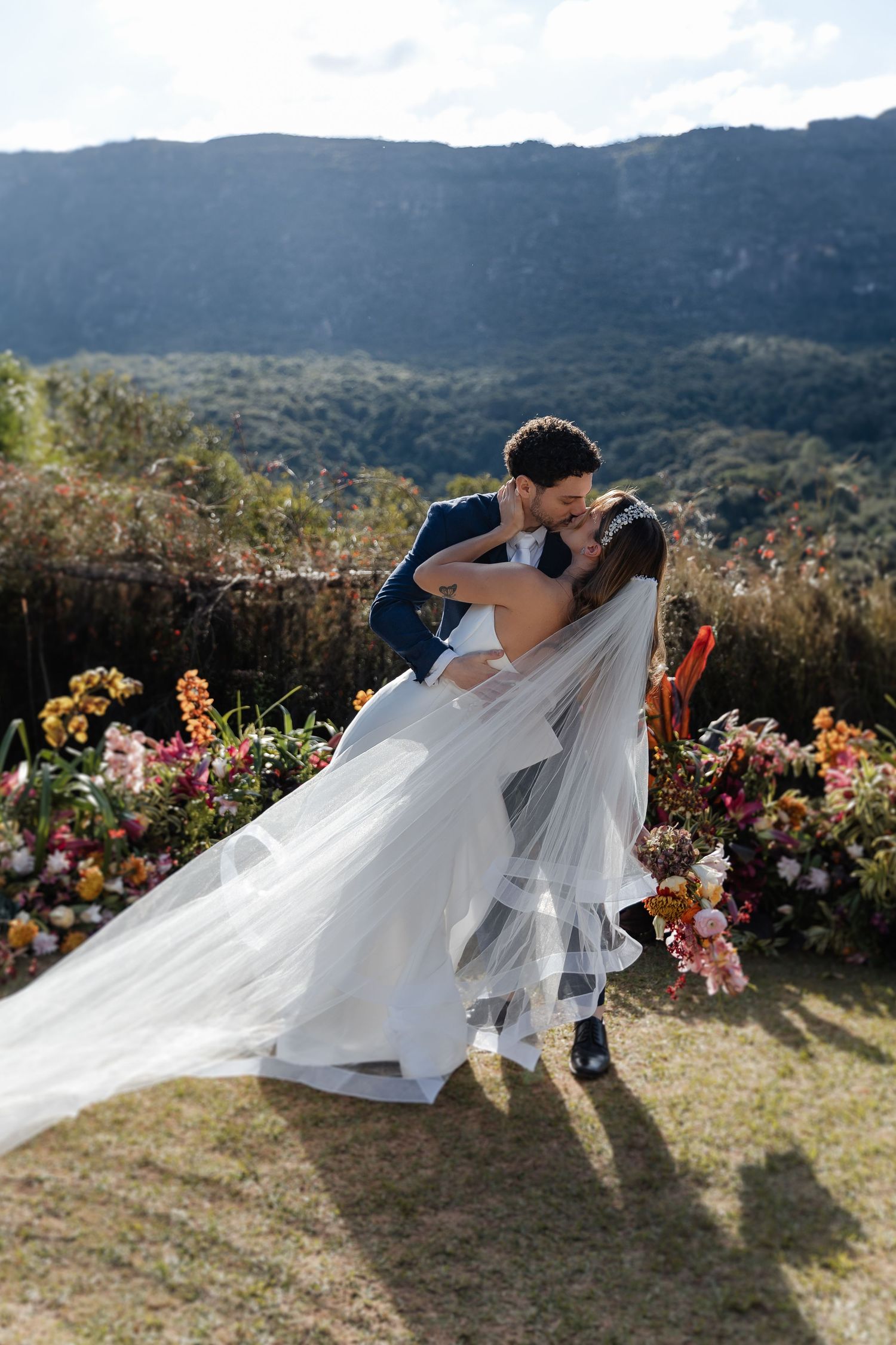 A couple in wedding attire share an intimate moment surrounded by flowers with mountain views in the background.
