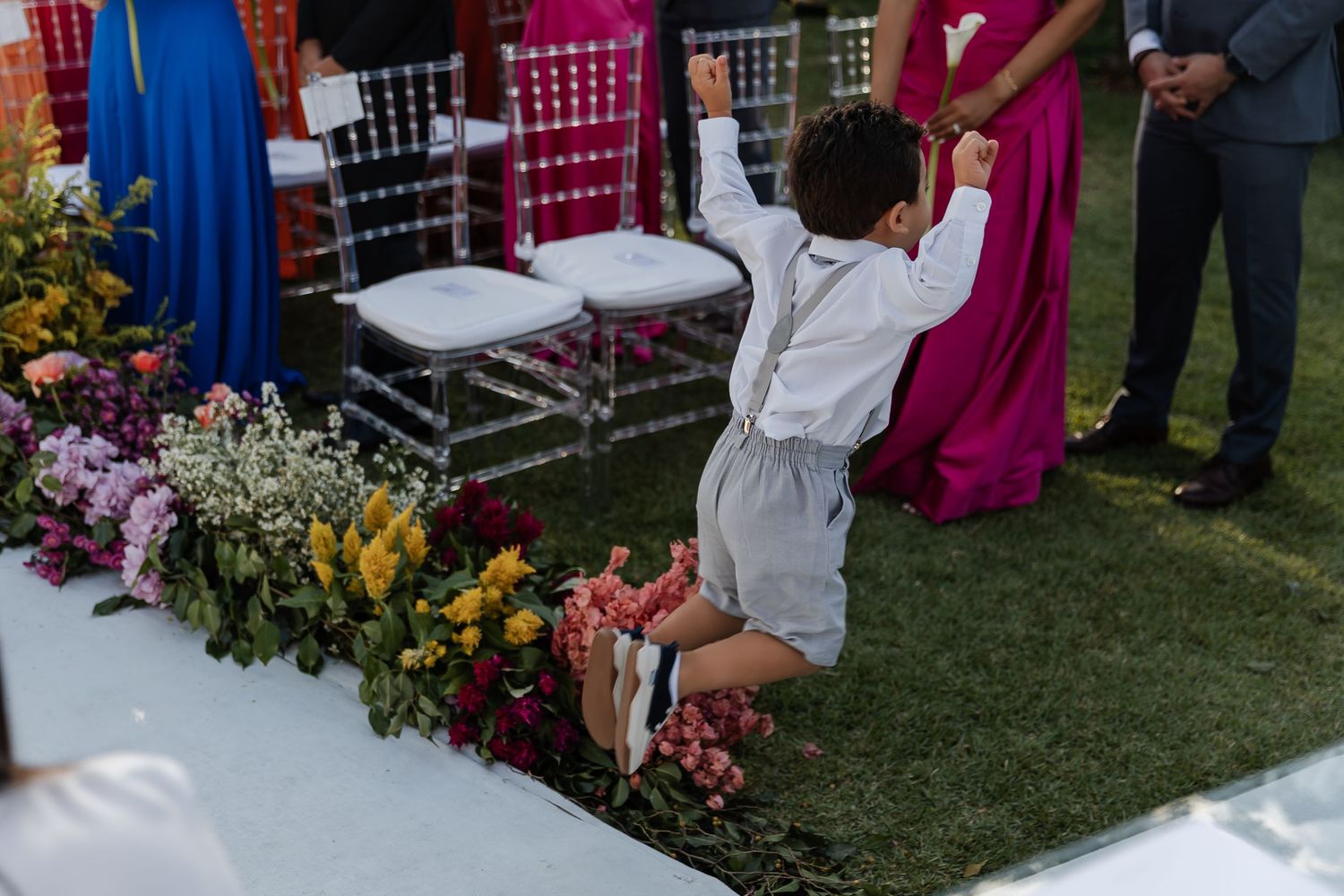 A child in white clothing jumps excitedly near colorful floral arrangements and chairs at an outdoor event.