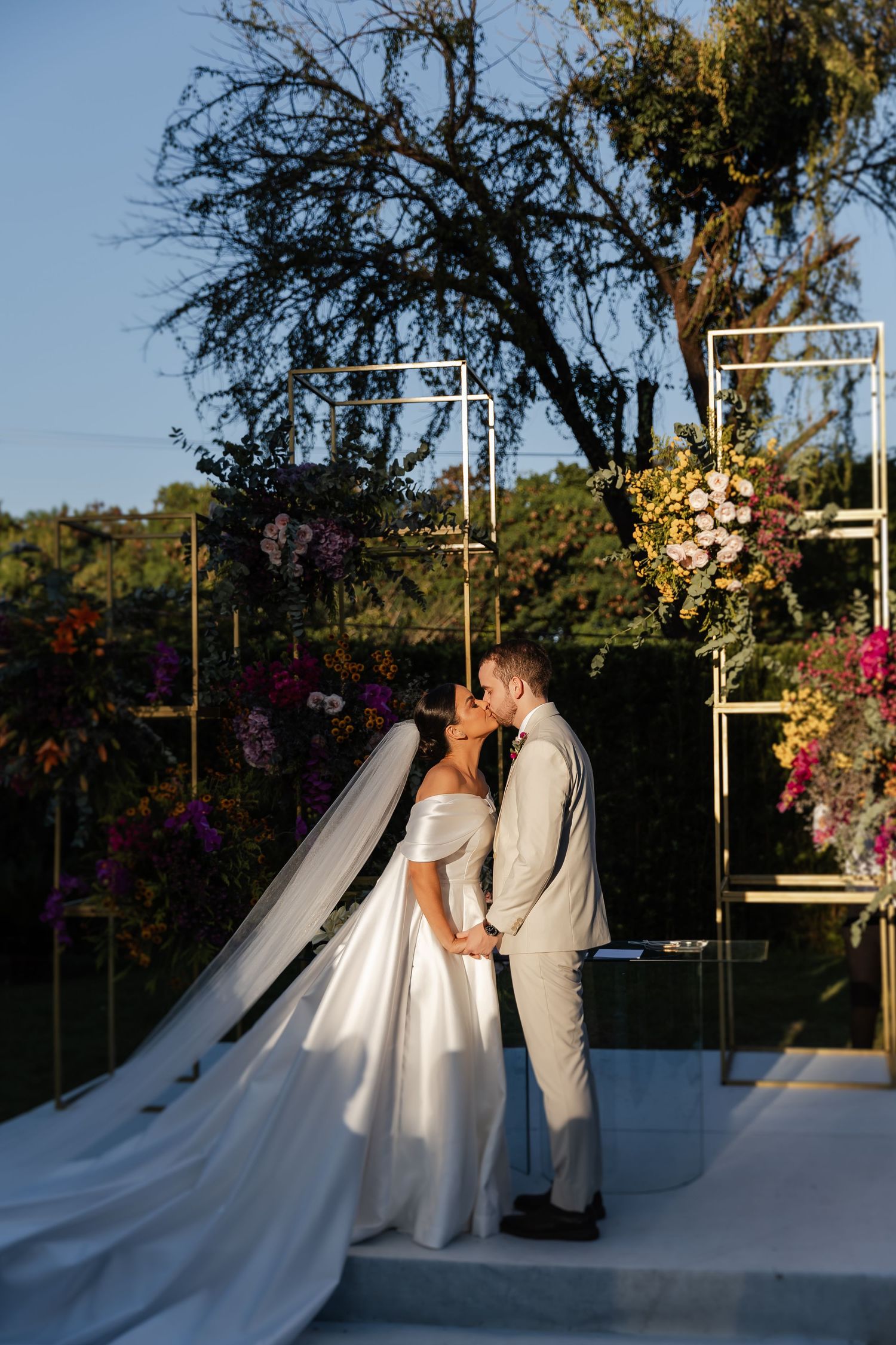 A couple shares an intimate moment under floral arches at an outdoor garden wedding ceremony at sunset.