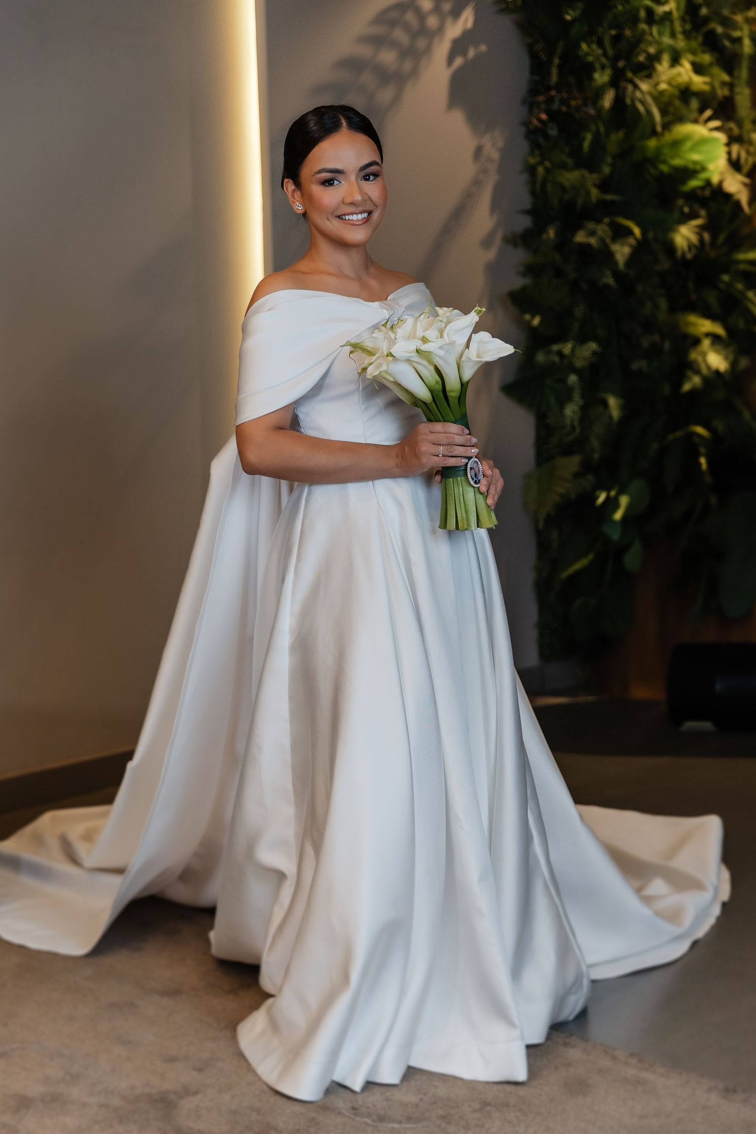 A bride in an elegant off-shoulder white satin wedding gown holds a bouquet of white calla lilies.