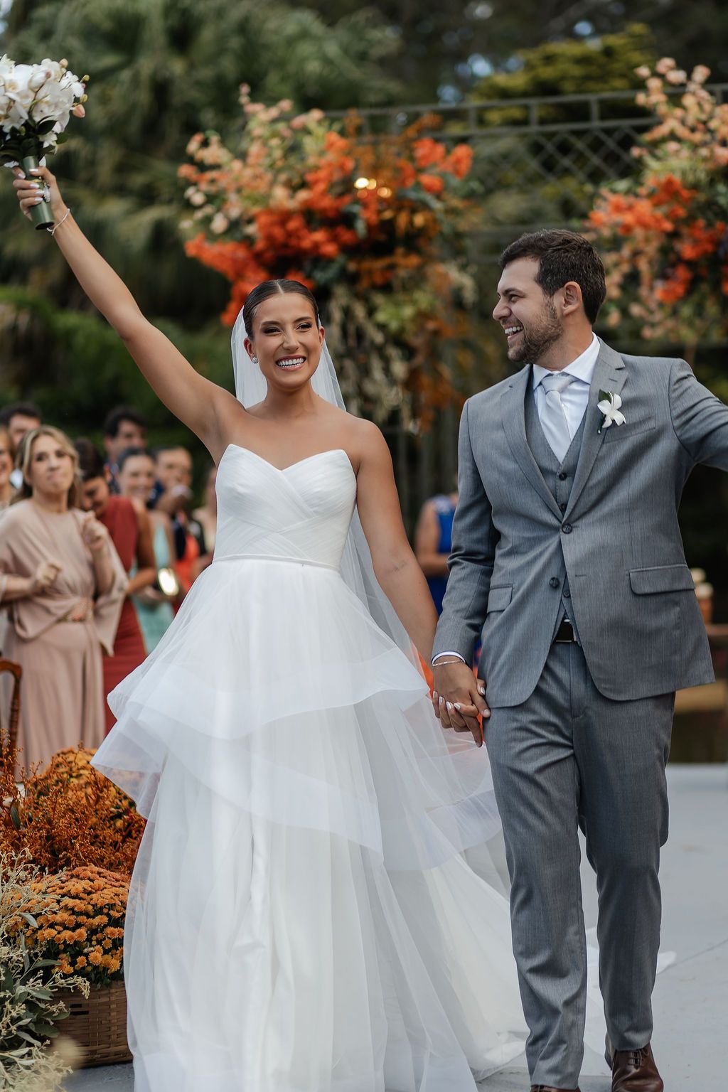 A joyous bride in a tiered white gown raises her bouquet while walking hand in hand with her groom in a gray suit.