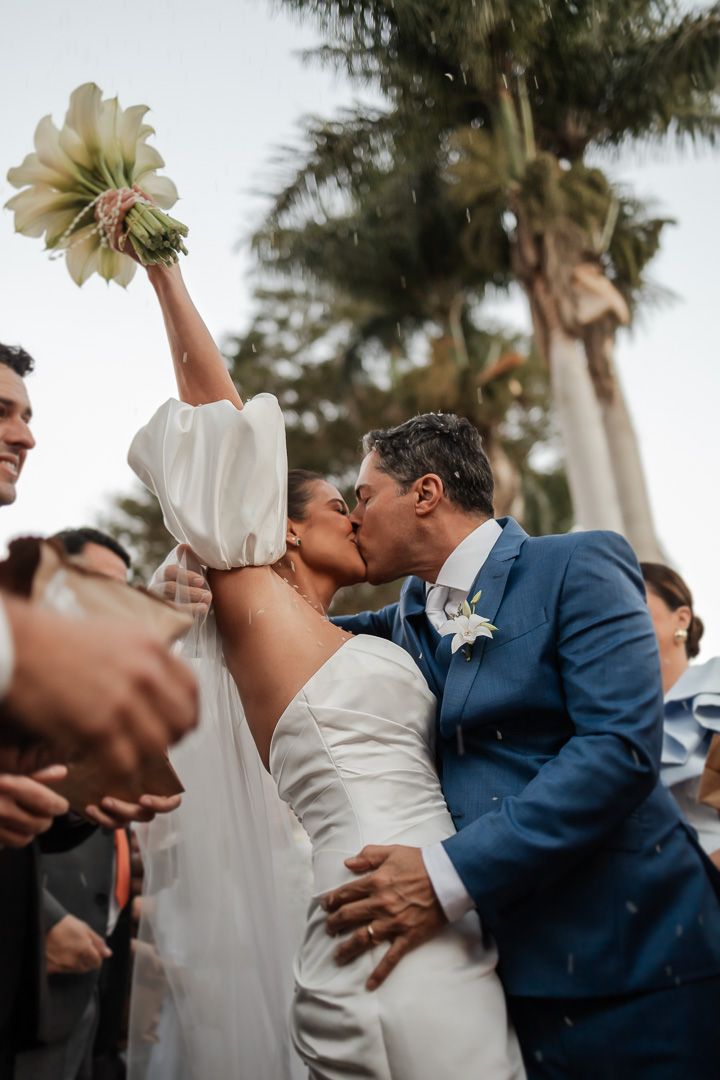 A joyful celebration at a tropical wedding as newlyweds share a kiss while guests cheer and palm trees sway in background.