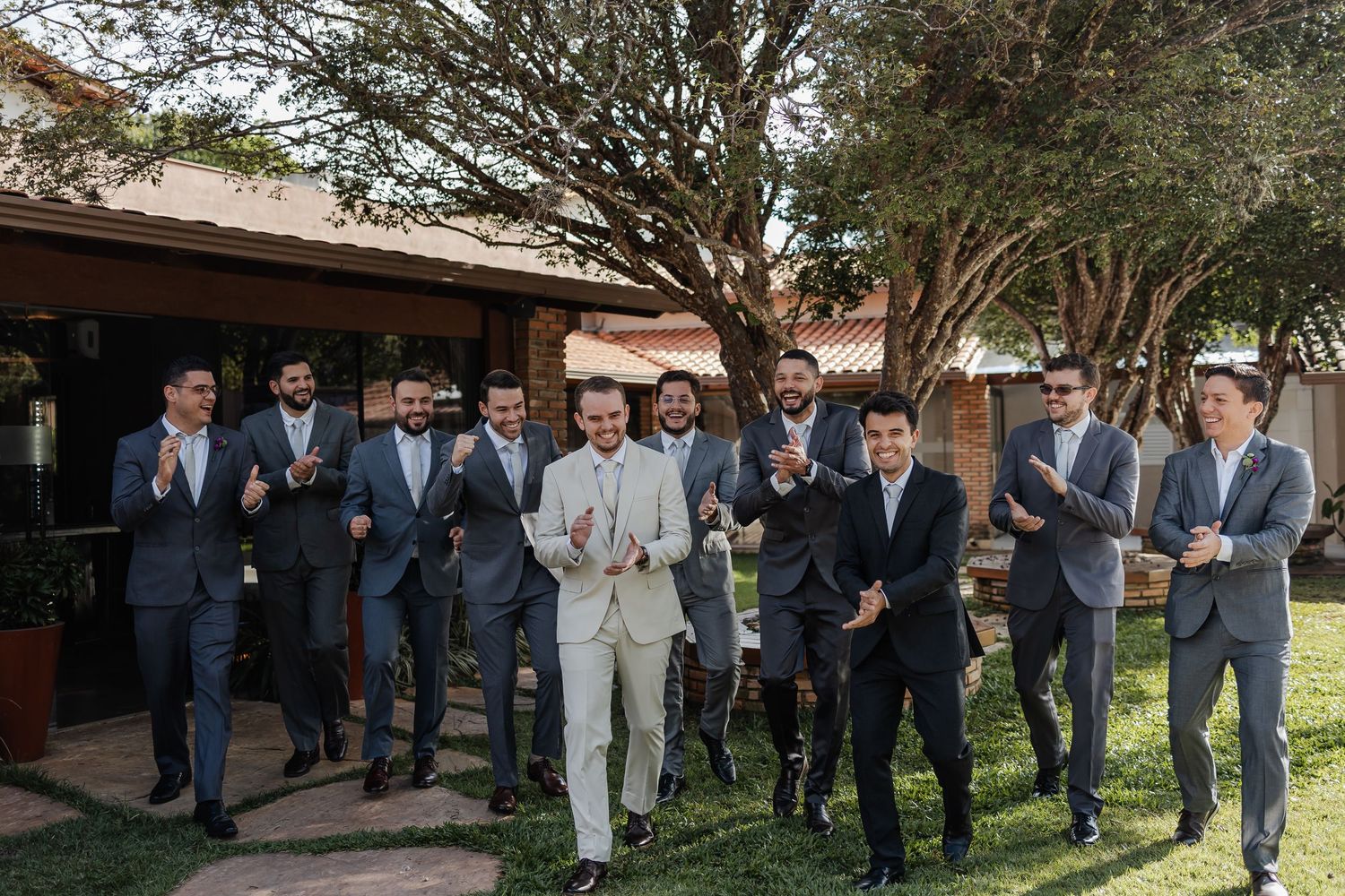 A wedding party in suits with varying shades of navy, gray, and beige stand together outdoors in front of trees.