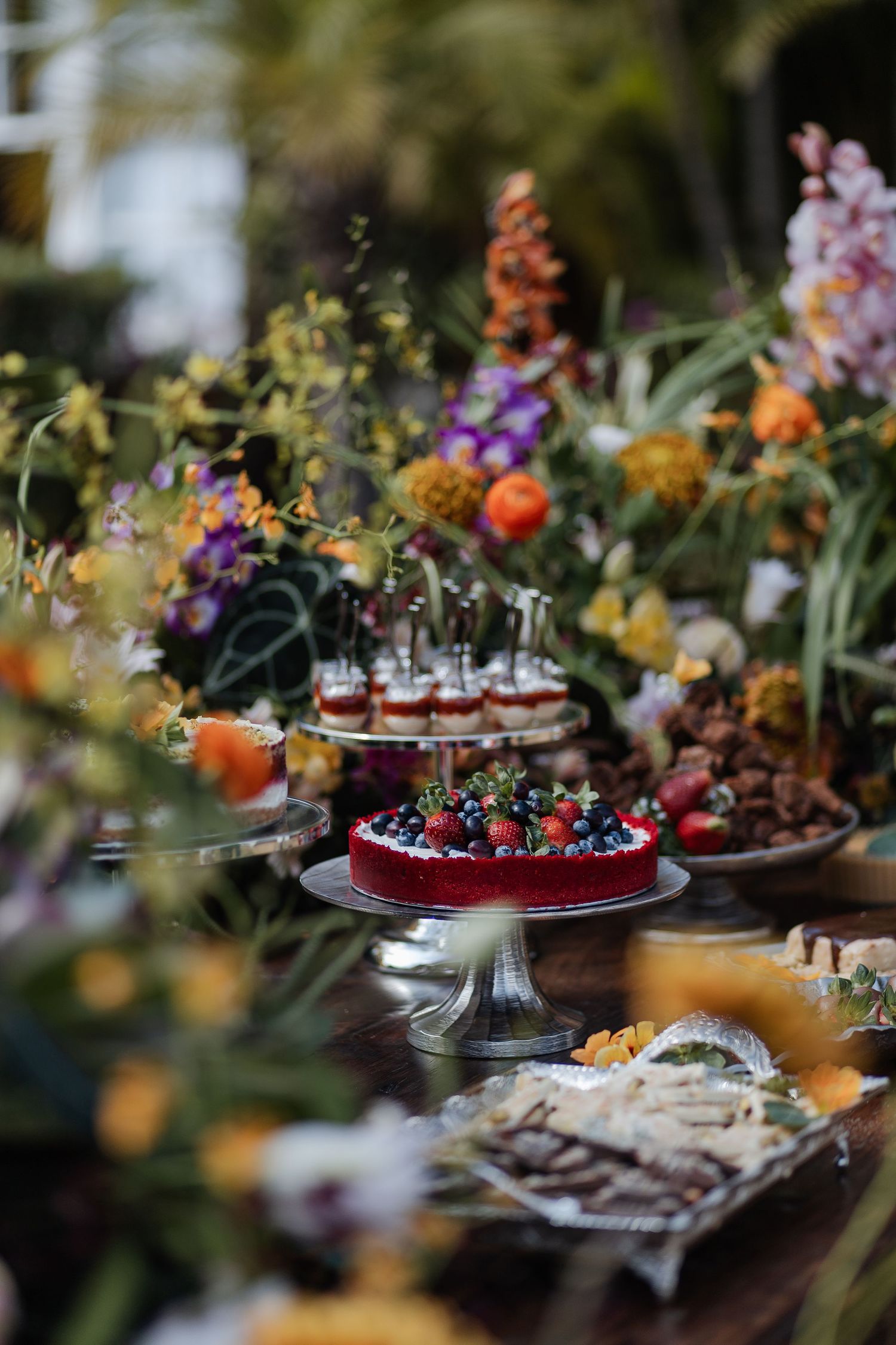 Elegant dessert table at outdoor reception with colorful cakes and treats surrounded by vibrant garden flowers.