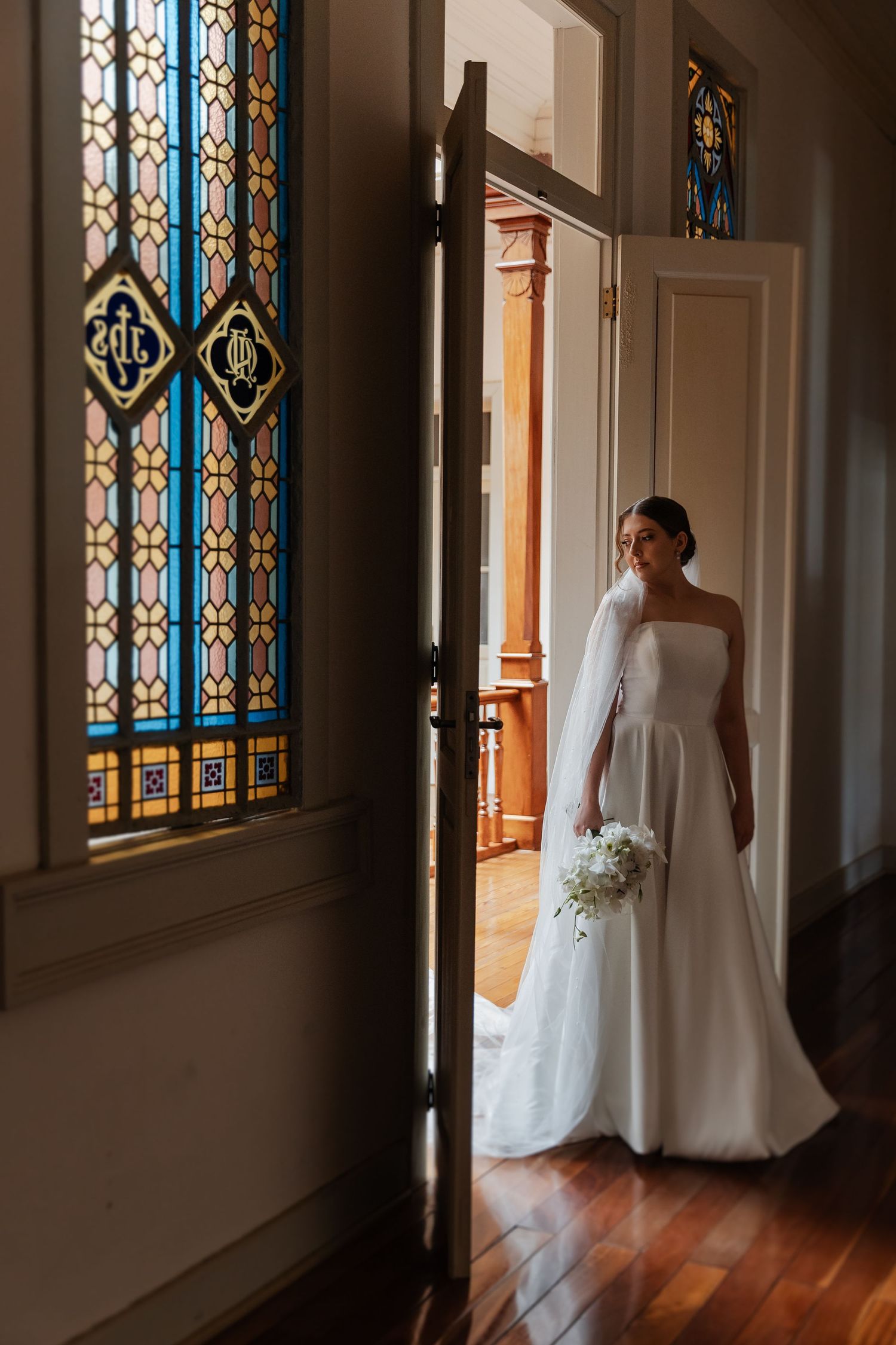 A bride in a white wedding gown stands silhouetted against stained glass windows in a church hallway.
