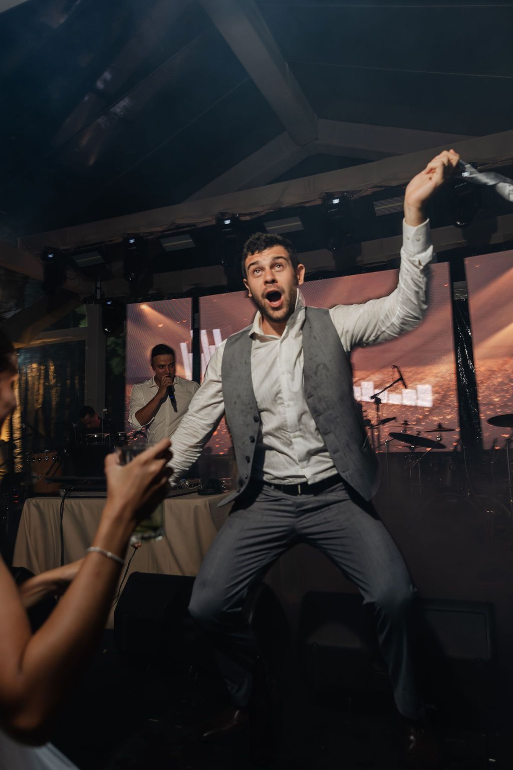 An energetic person in a grey vest and white shirt jumps excitedly while dancing at a dimly lit reception venue.