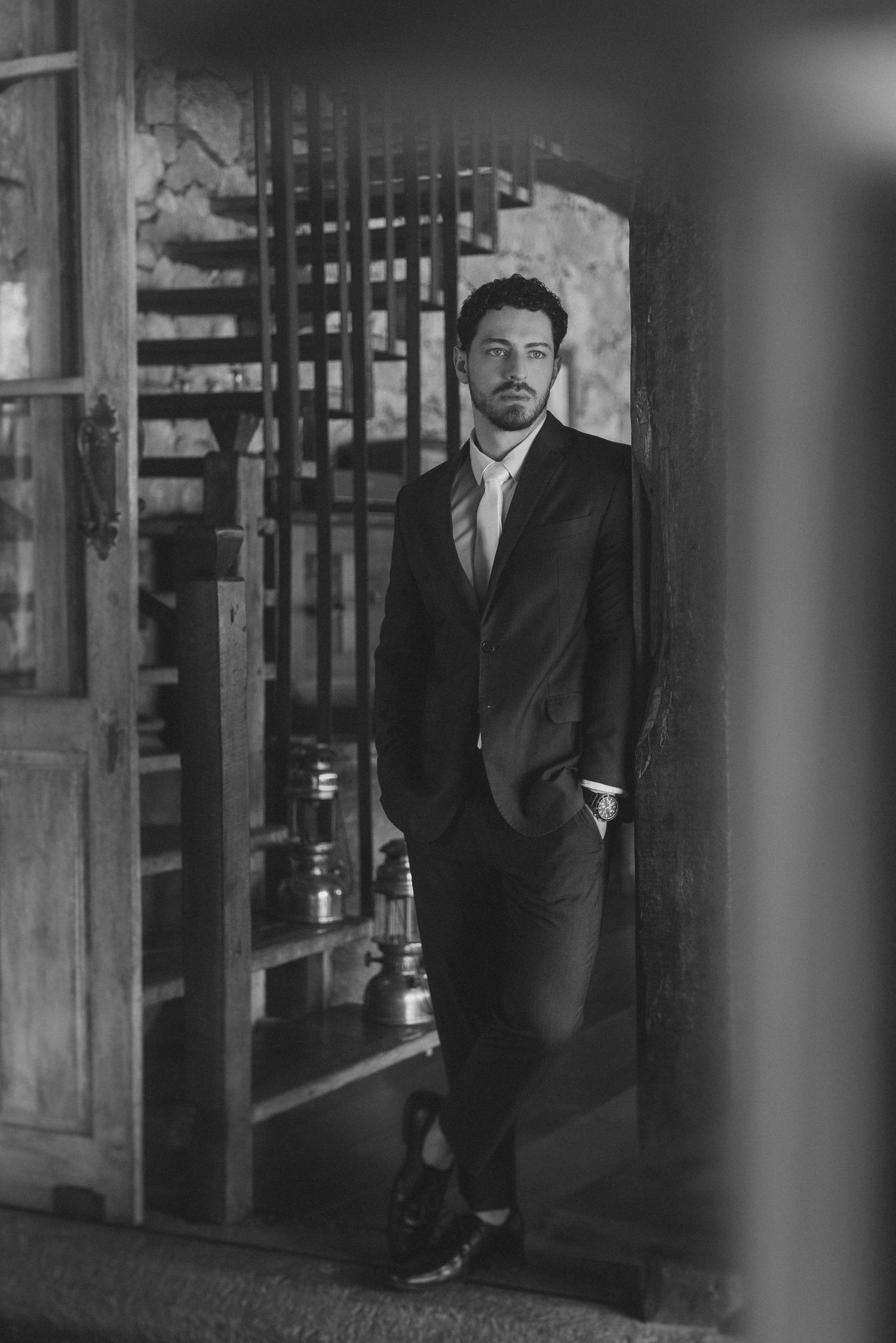 A black and white portrait of someone in a formal suit standing against a rustic doorway with stairs visible.