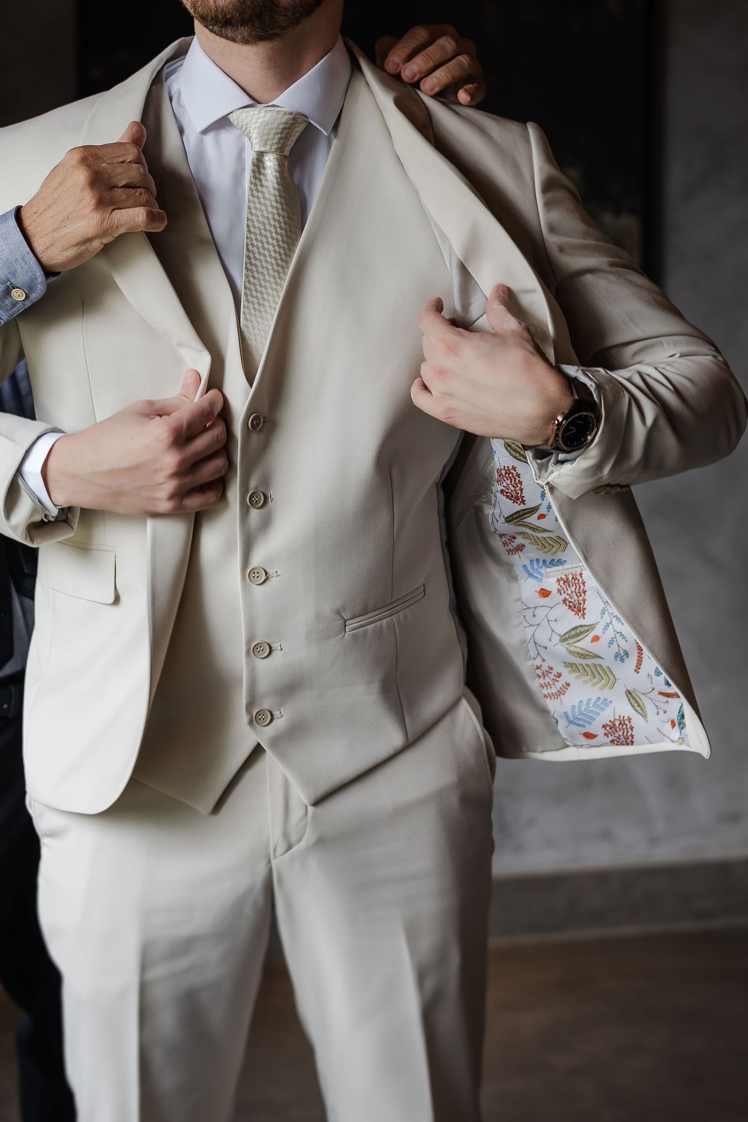 Close-up of a cream-colored three-piece suit with patterned lining and silver tie showing elegant formal menswear details.