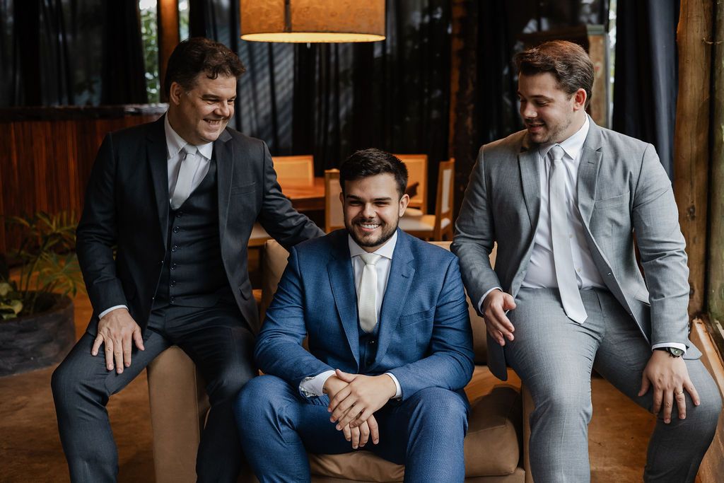 Fotógrafo de casamento em SP,Three groomsmen in formal suits relaxing and laughing together at an indoor venue before a wedding celebration.