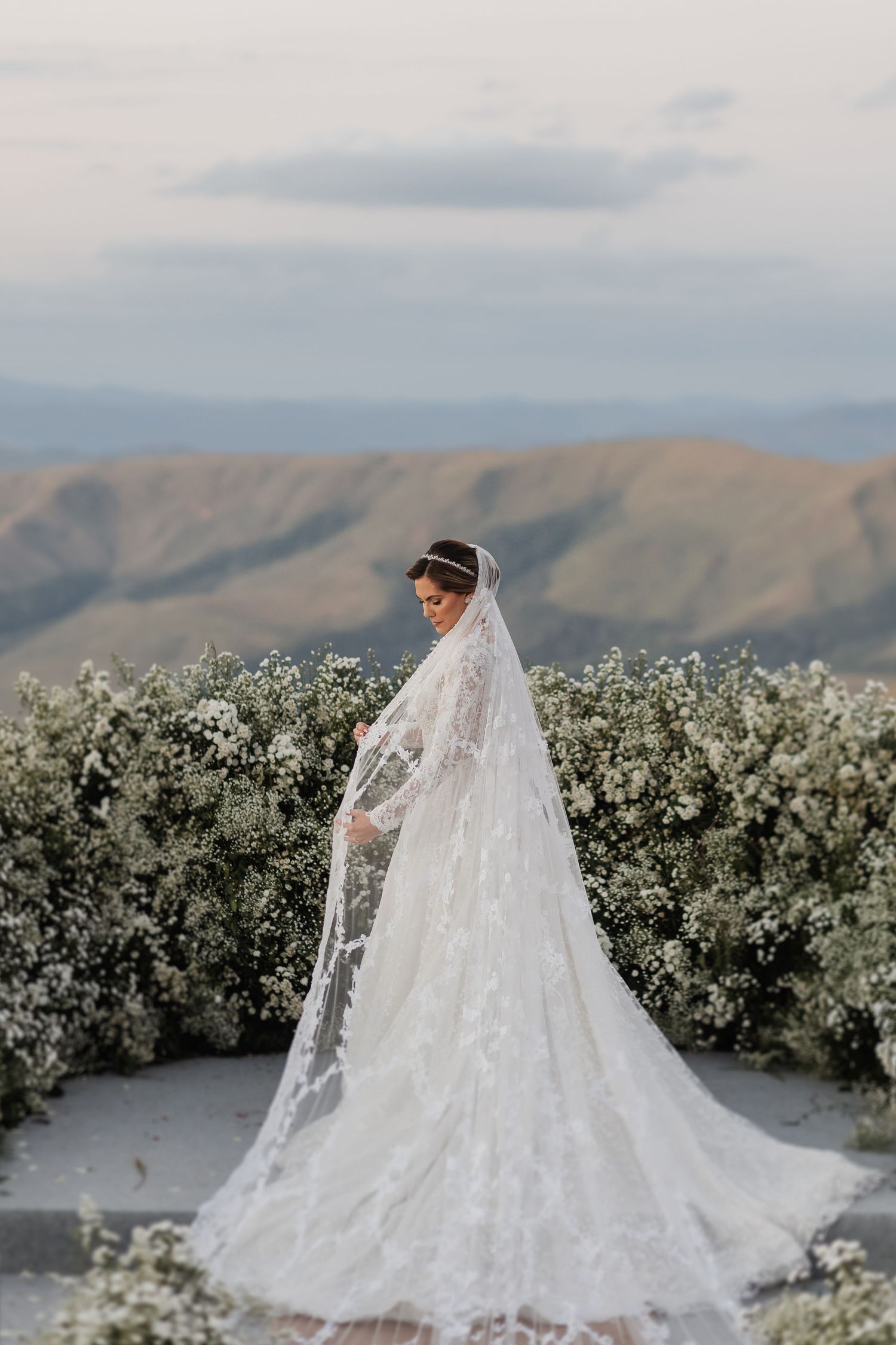 Elegant bride in flowing lace veil poses against mountain landscape with white floral backdrop on wedding day.