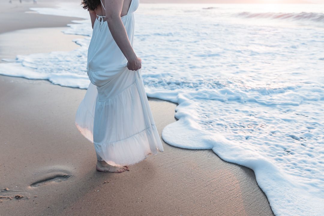Woman in flowing white dress walking barefoot on sandy beach with gentle ocean waves at sunset.