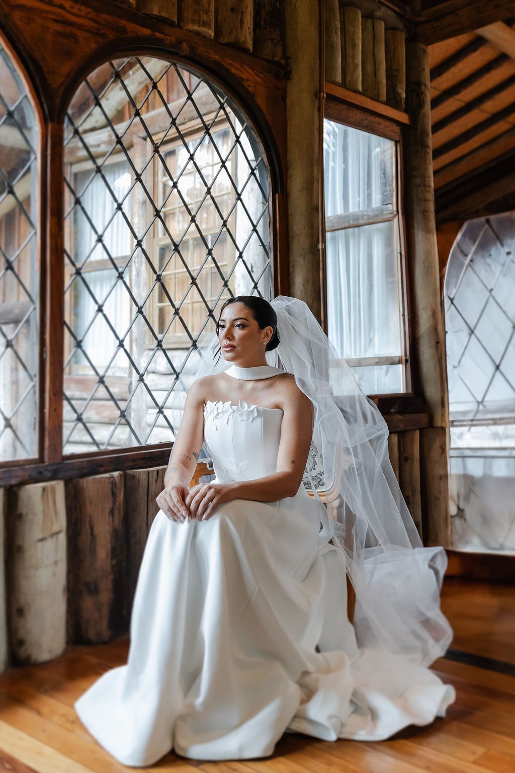 Fotógrafo de casamento em SP,A bride in a crisp white strapless gown and flowing veil poses by a window with diamond-patterned leaded glass.