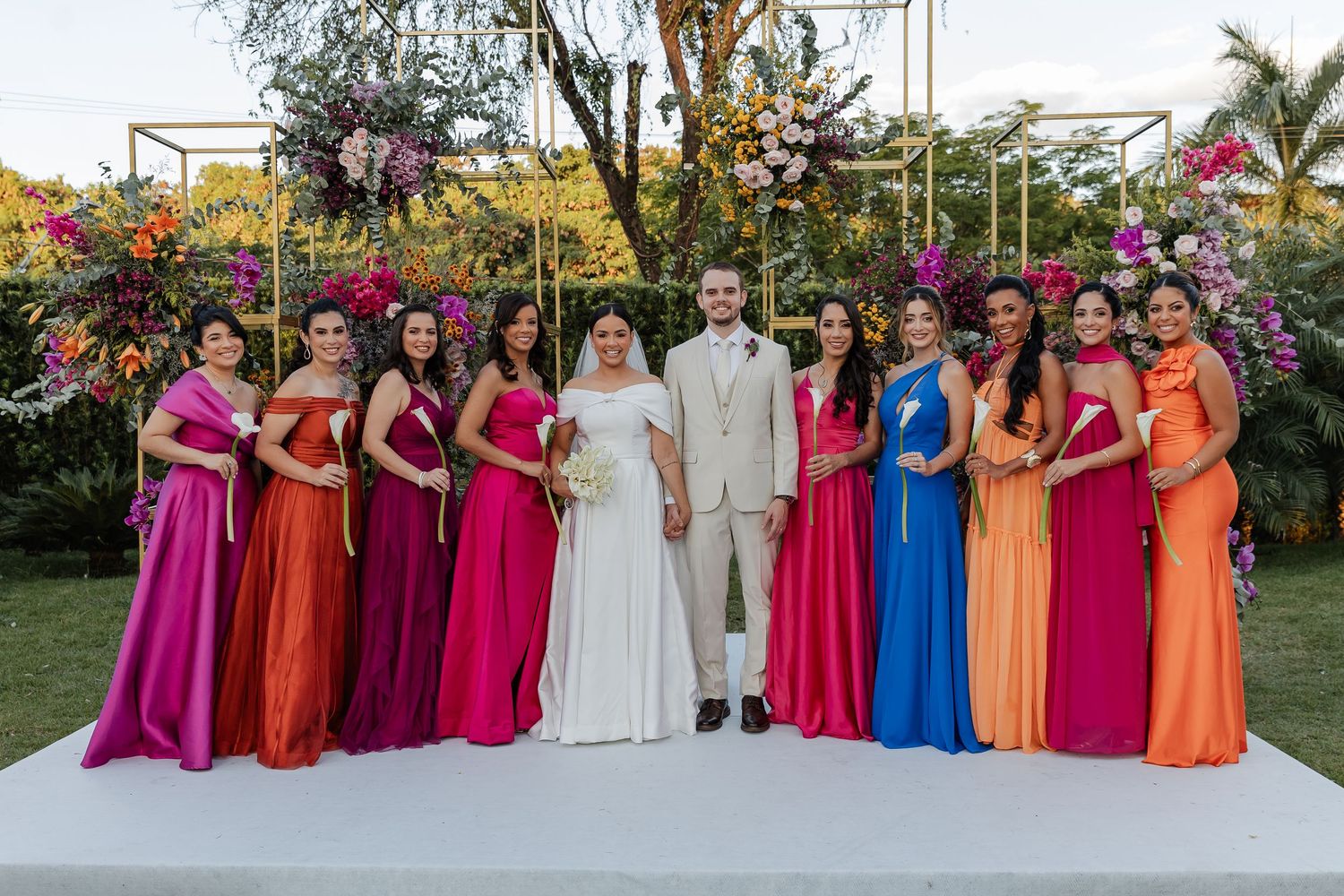 Wedding party stands in colorful rainbow-ordered formal attire on a white platform in a garden setting.