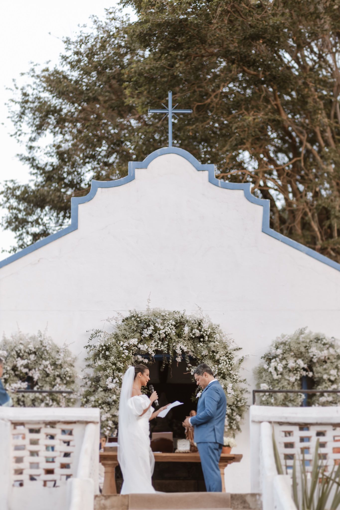 A wedding ceremony takes place at a charming white chapel with a blue cross and floral arch decorations.