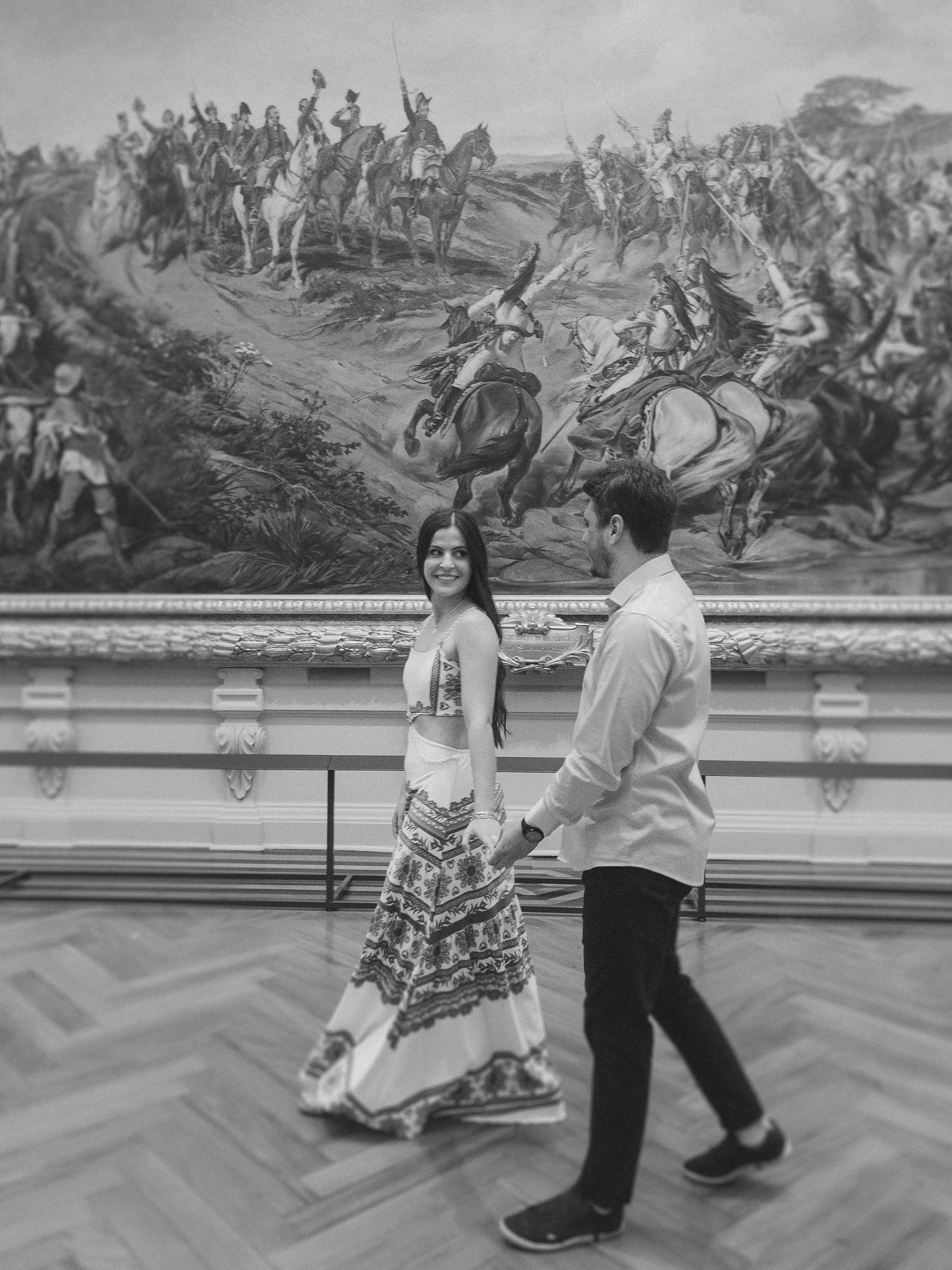 Two visitors walk past a large historical battle scene painting displayed in an art gallery with herringbone wood flooring.