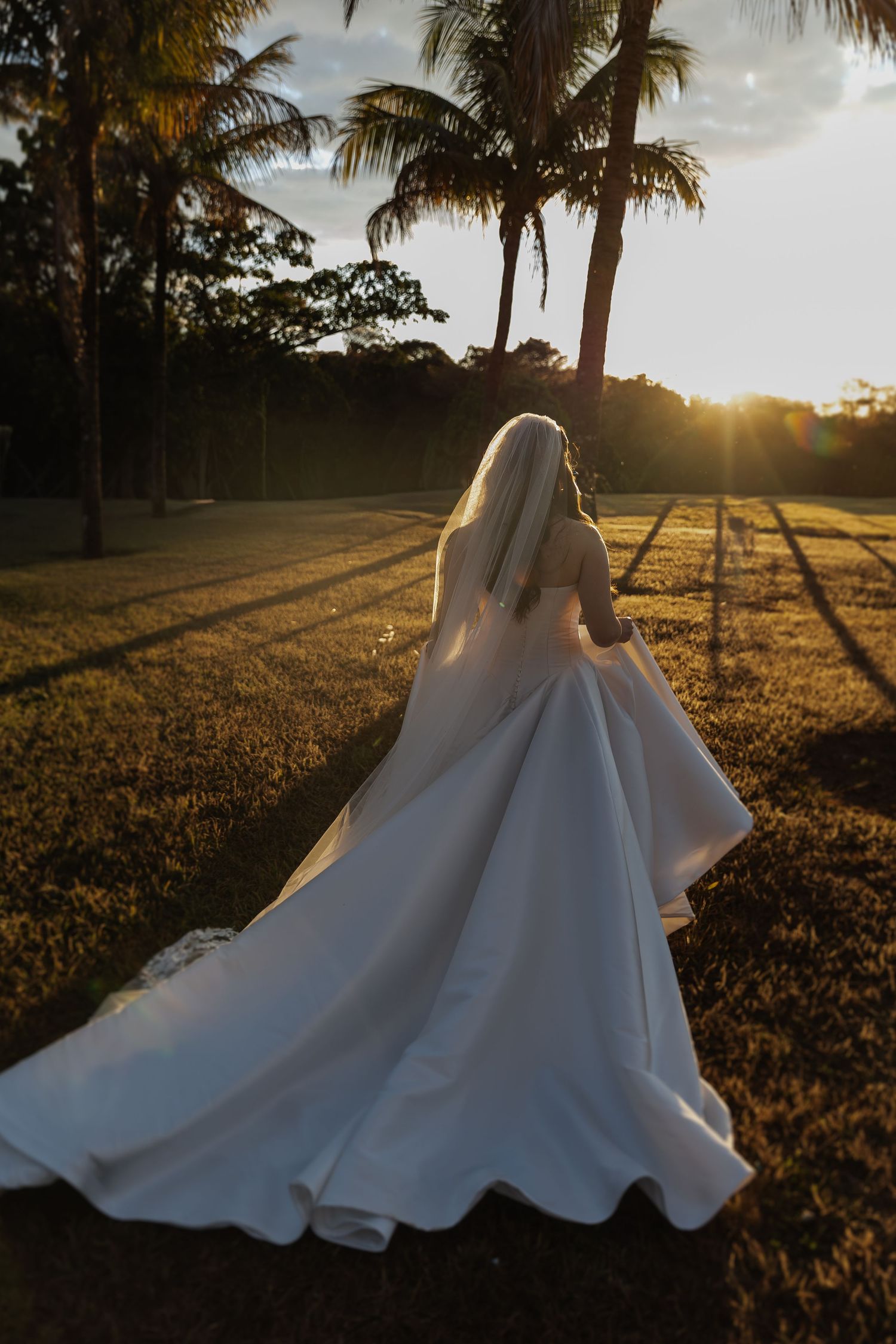 A flowing white wedding dress billows in the golden sunlight as palm trees sway in the background at sunset.